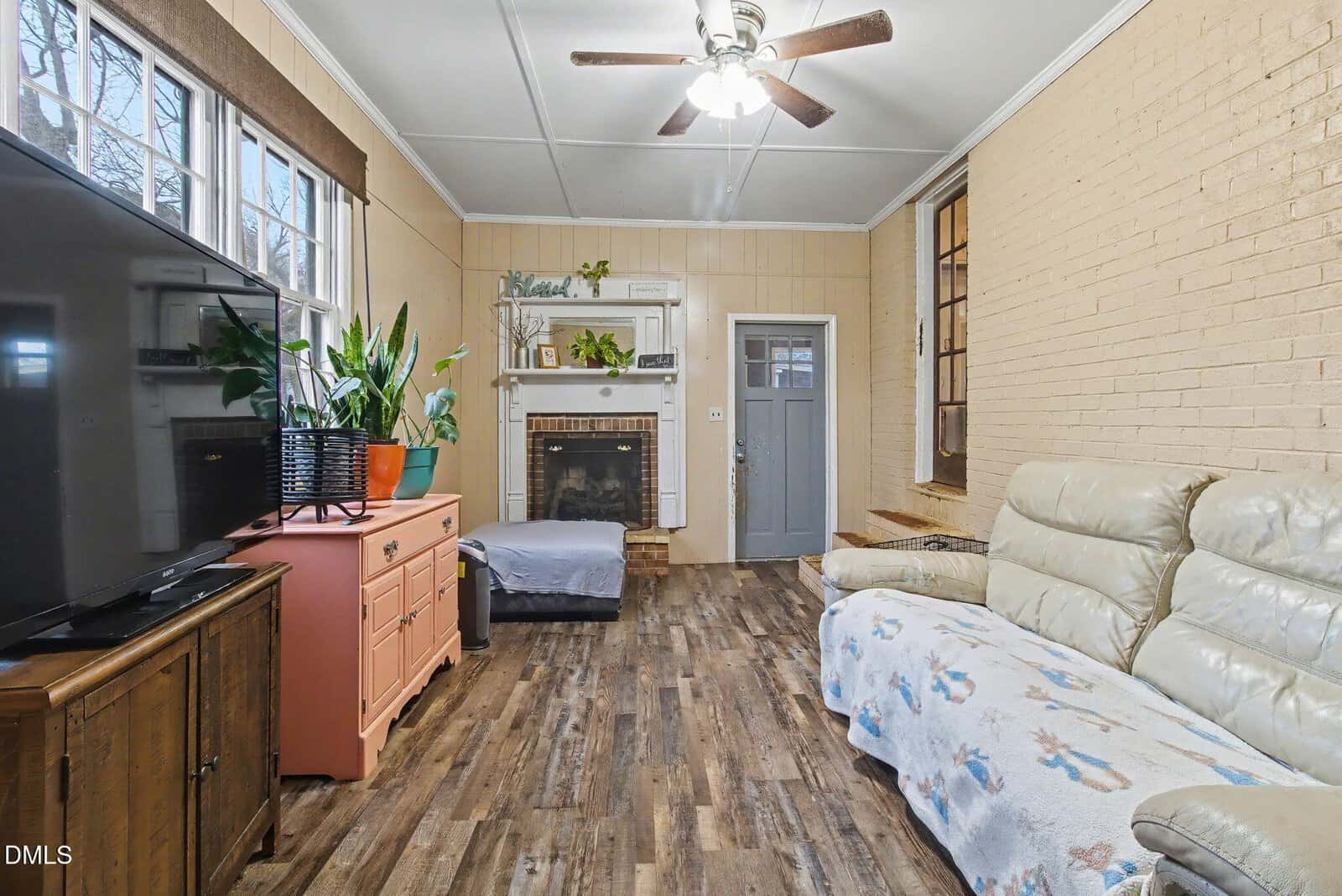 Cozy living room with wood-style flooring, a light-colored leather sofa, a fireplace, large TV, potted plants, a coral cabinet, and a ceiling fan; neutral walls and a gray door add to the warm ambiance.