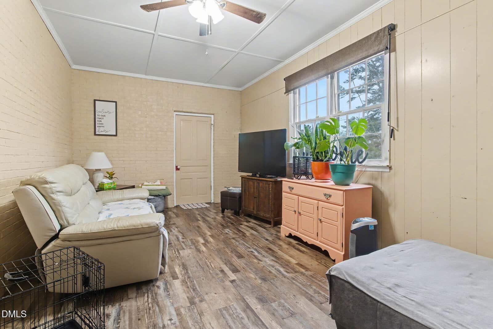 Cozy living room with tan walls, wood floor, beige sofa, TV on wooden stand, orange cabinet with potted plants, window with brown shade, ceiling fan, framed art, and small black pet crate in the corner.