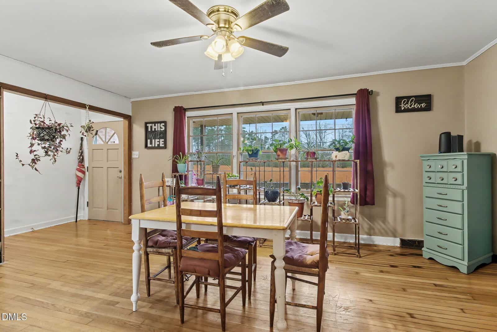 Dining room with a yellow table, five mismatched chairs, hardwood floor, ceiling fan, and large windows with plants. Wall art says “THIS IS LIFE” and “Believe.” Green dresser and hanging plant also visible.