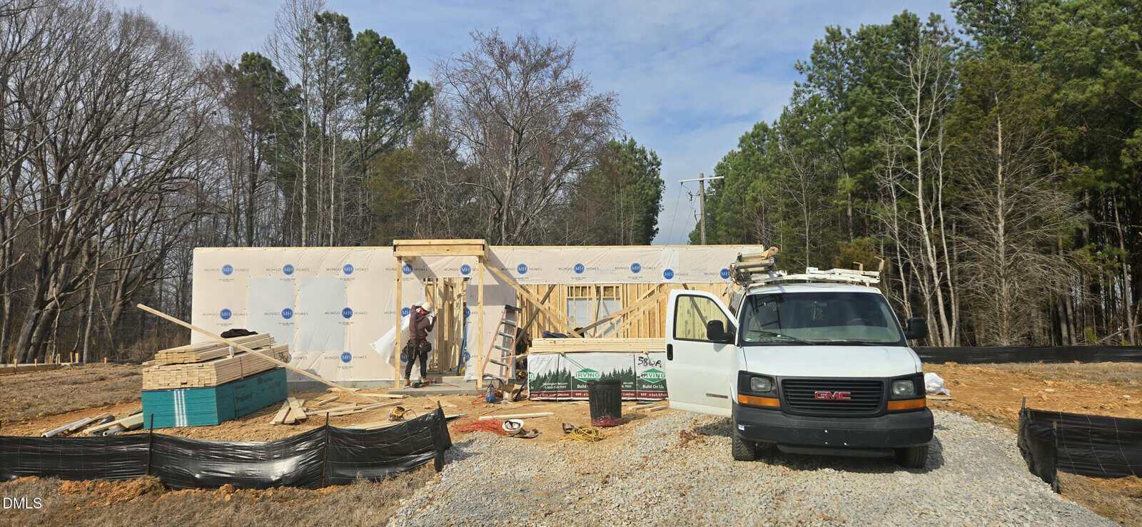 A house under construction with exposed framing and wrapped walls, two workers near the entrance, a white GMC van parked in front, building materials scattered, and trees in the background.