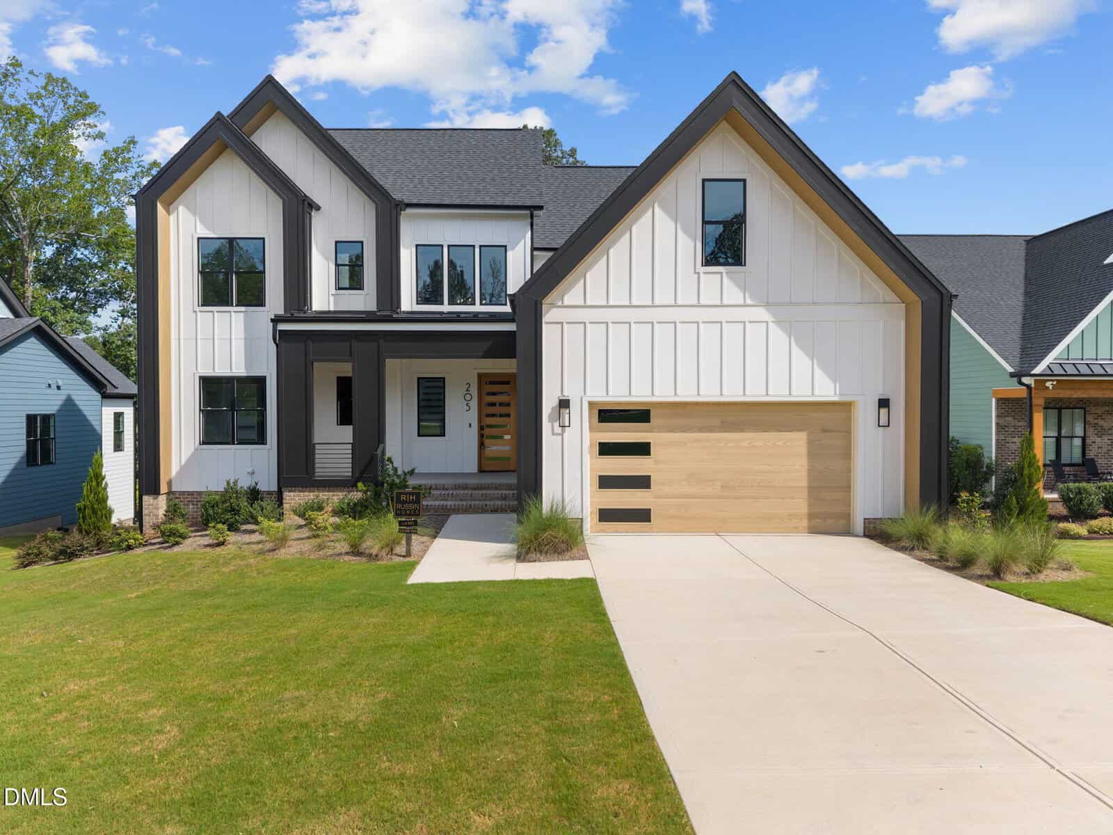 Modern two-story house with black roof, white and wood siding, large windows, and a double garage with wood doors. Manicured lawn and driveway in front, with blue sky and clouds above.