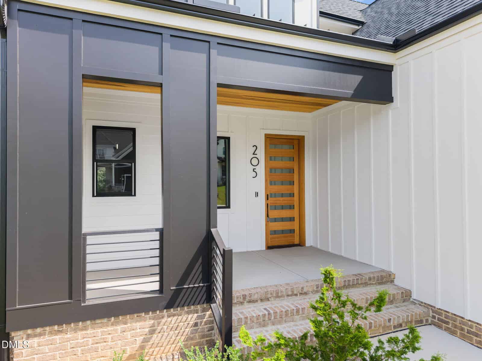 Modern house entryway featuring a wooden front door with horizontal glass panels, white vertical siding, black accents, brick steps, and a metal railing. The house number "205" is displayed next to the door.