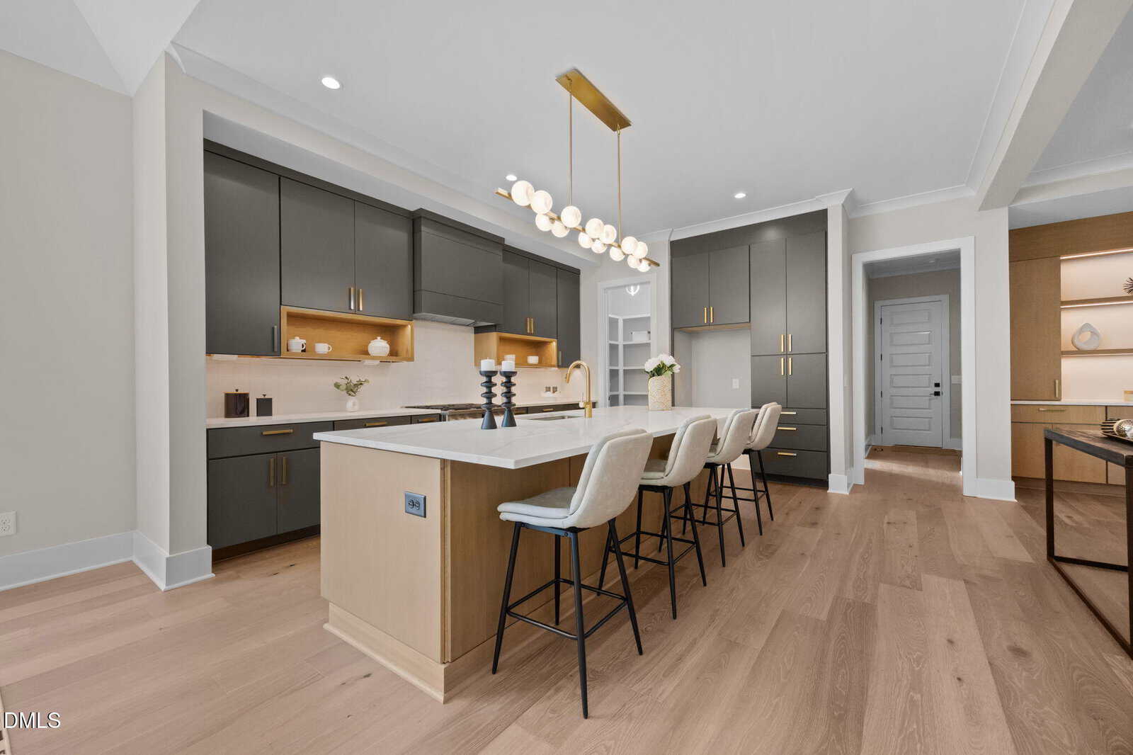 Modern kitchen with gray cabinets, gold hardware, a large island with white countertop, four white barstools, wooden floors, and a contemporary gold light fixture above the island.
