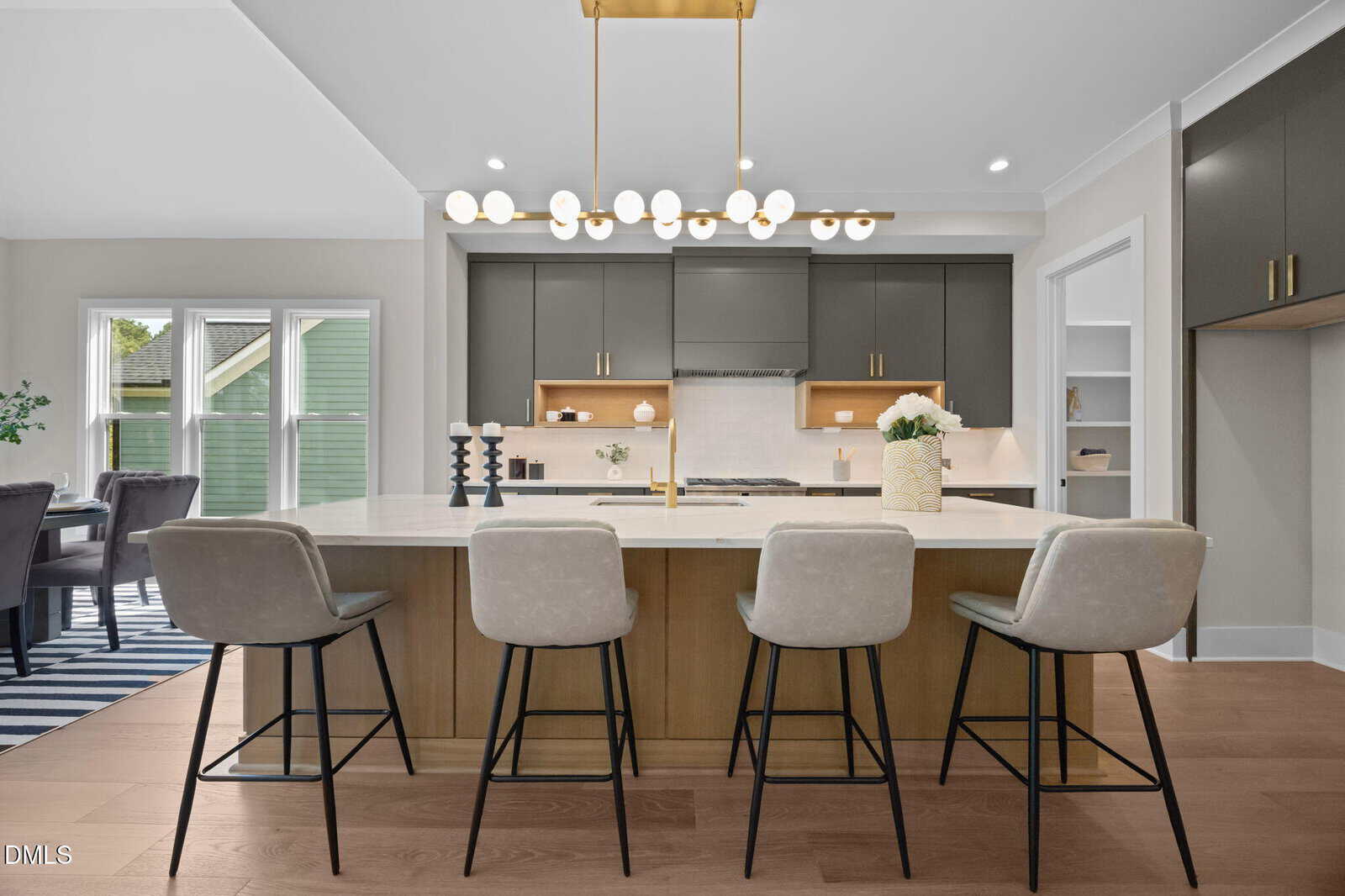 Modern kitchen with a large island, four upholstered bar stools, dark cabinets, open shelves, a white countertop, pendant lighting, and a dining area visible to the left by a set of windows.