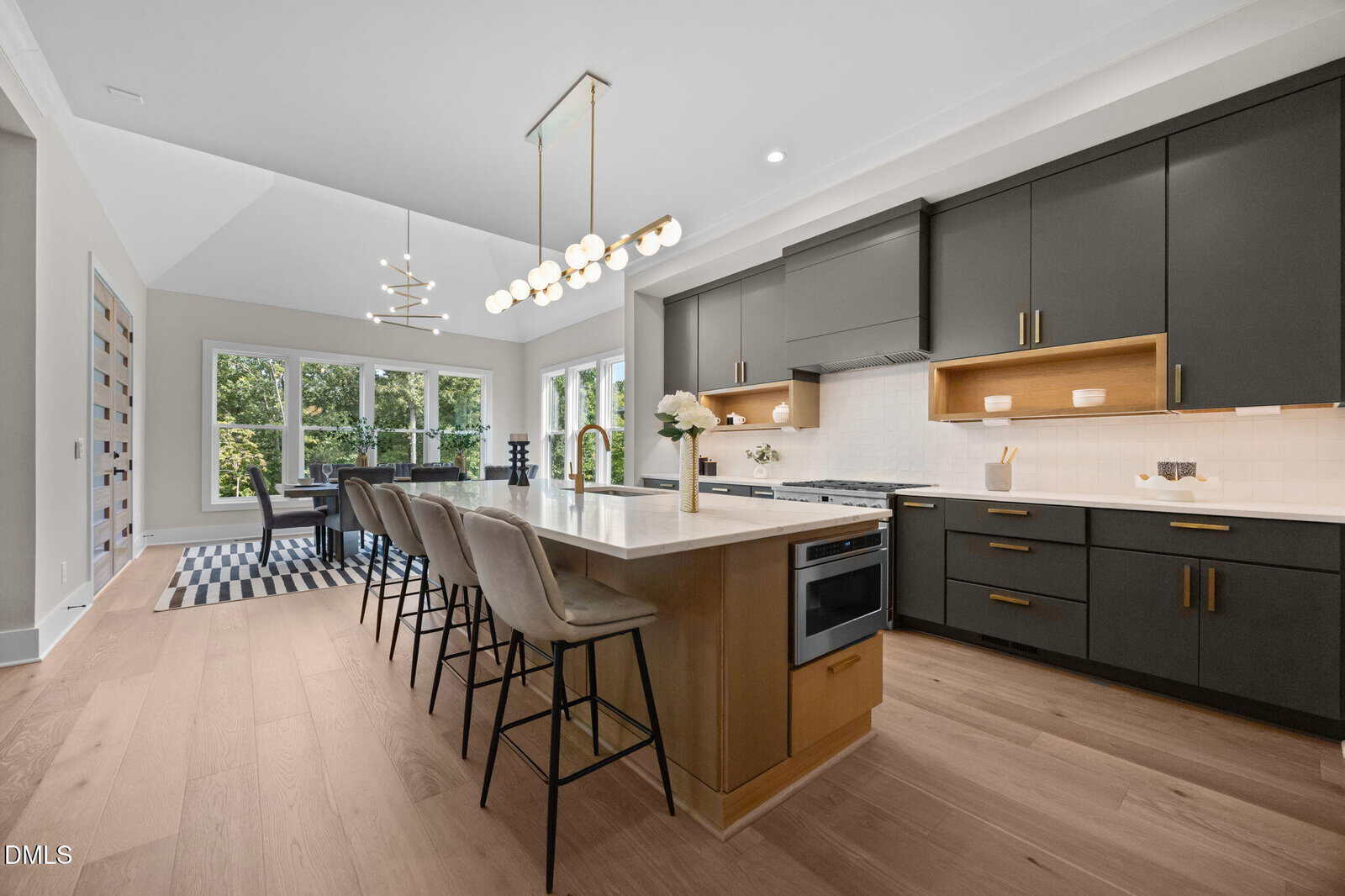 Modern kitchen with black cabinets, wood accents, and a large island with barstools. Pendant lights hang above the island. The dining area with large windows is visible in the background.