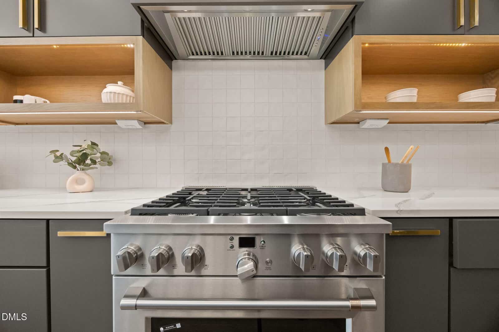 Modern kitchen with a stainless steel gas stove, white tile backsplash, dark cabinets, open shelves holding dishes, and a small plant on the left counter.