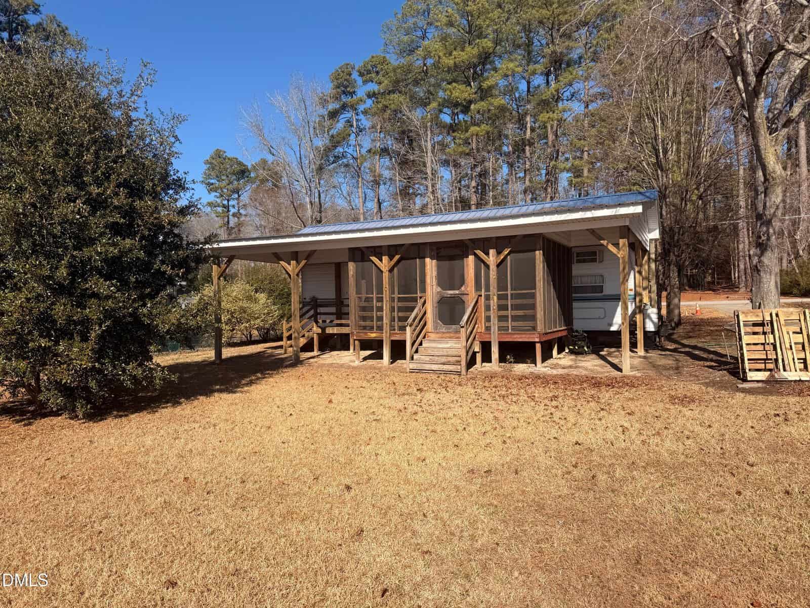 A small cabin with a covered porch sits in a grassy yard at 1561 Sagamore Drive, Louisburg, surrounded by trees. Steps lead up to the porch, supported by wooden posts, and pallets are stacked to the right of the building.