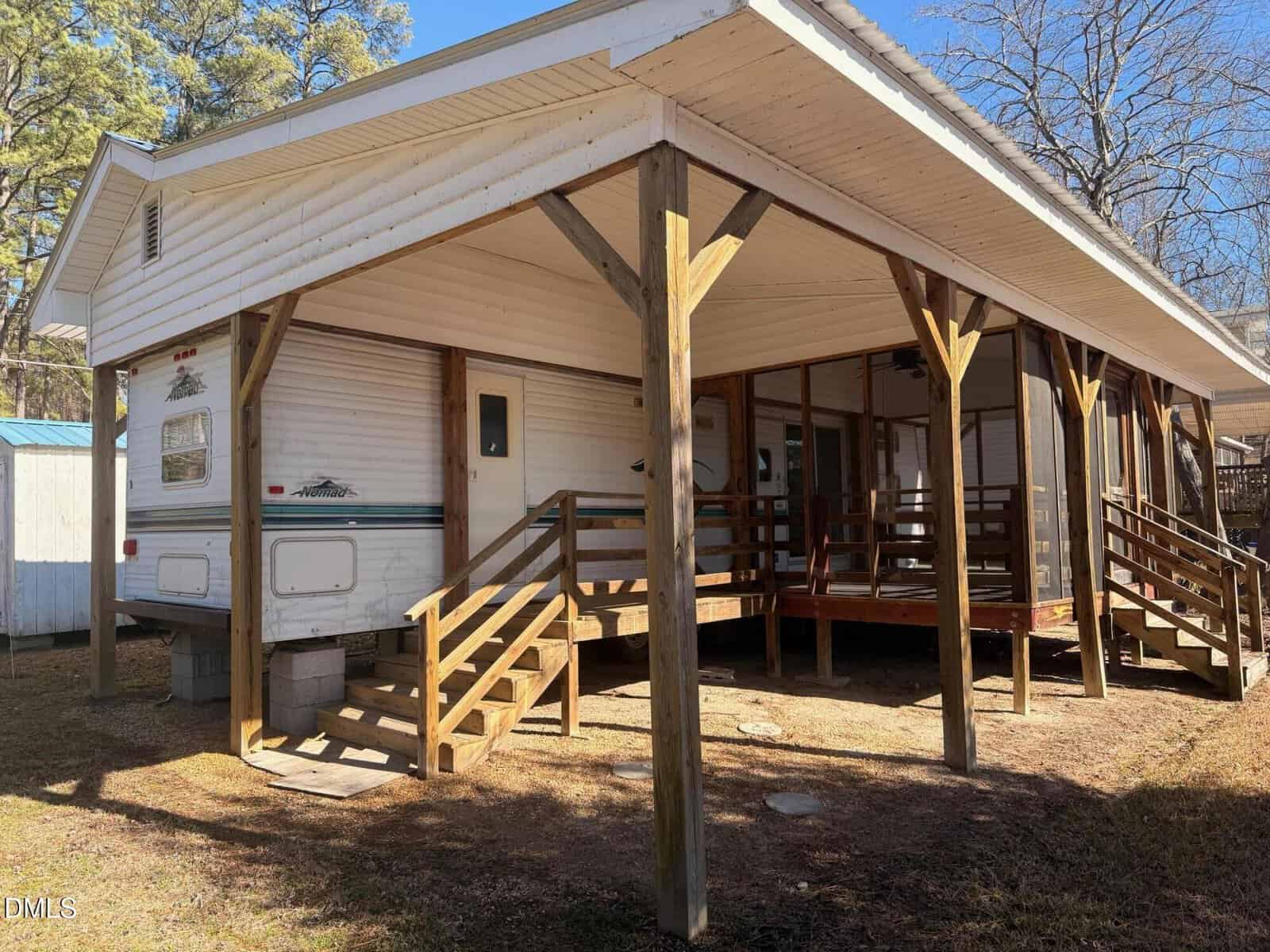 A white travel trailer is parked under a wooden, roofed structure with two sets of stairs leading to a screened porch area. Shadows fall on the ground, and tall trees are visible in the background.