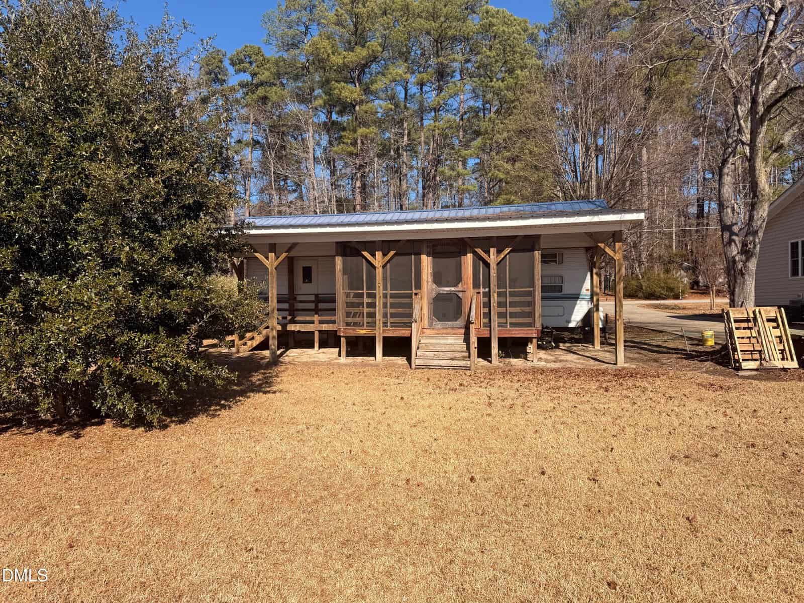 A small, single-story house with a metal roof and a screened front porch sits on a dry, grassy lawn. Tall trees are in the background, and a pile of wooden pallets is on the right side near another building.