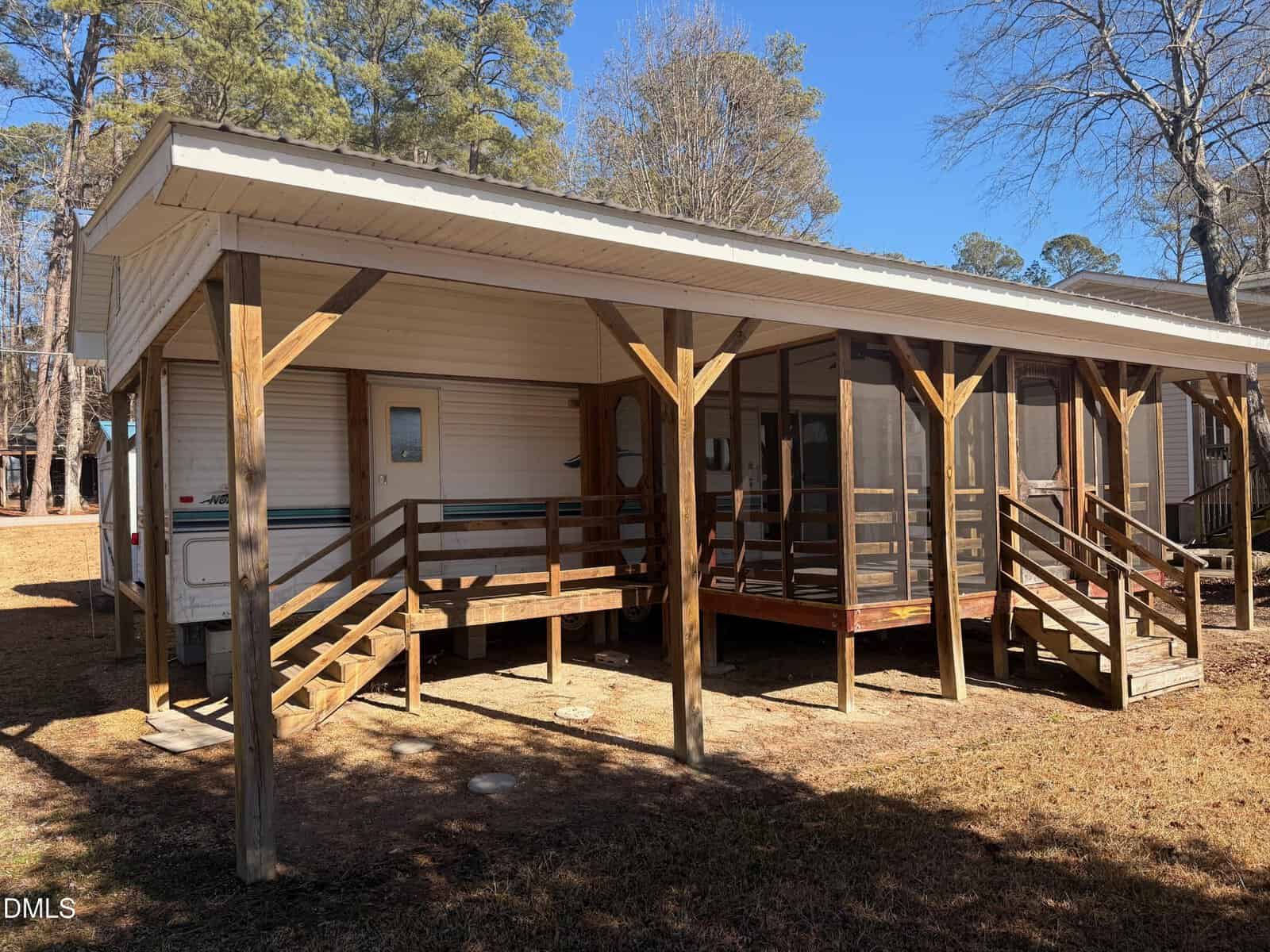 A white mobile home with a wooden porch and ramp under a slanted roof, surrounded by trees and grass on a sunny day. Steps lead up to the porch, and screened windows are visible on the right side.
