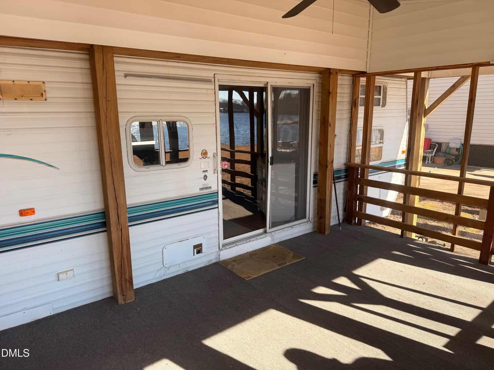 Enclosed porch with carpeted floor and wooden railing, overlooking a body of water. A sliding glass door leads into a camper or RV with white siding and blue-green stripes. Sunlight casts shadows on the porch.