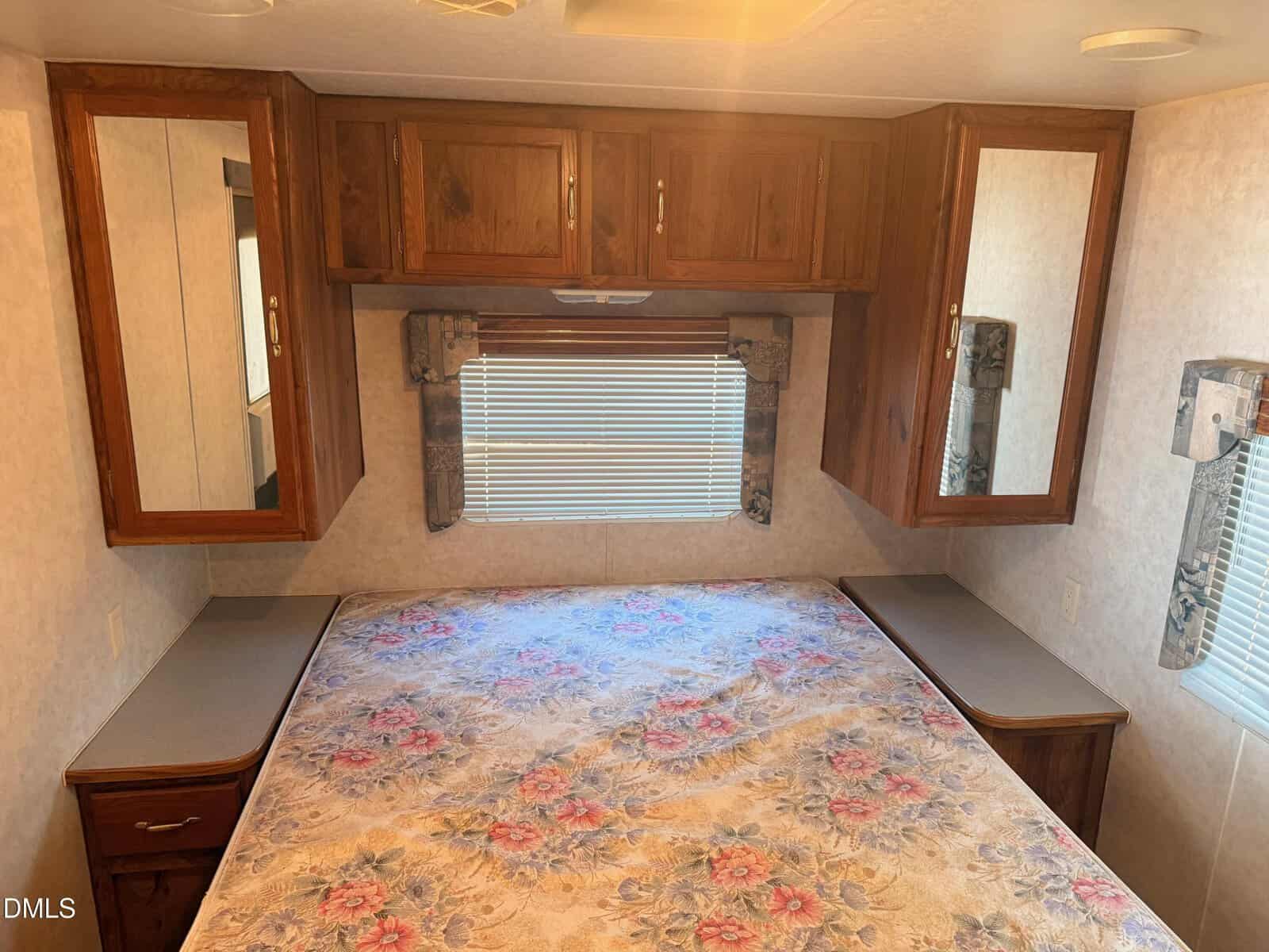 A small bedroom with a floral-patterned bed, wood cabinets above, mirrors on both sides, side tables, and a window with blinds and valances. The decor is simple and traditional.