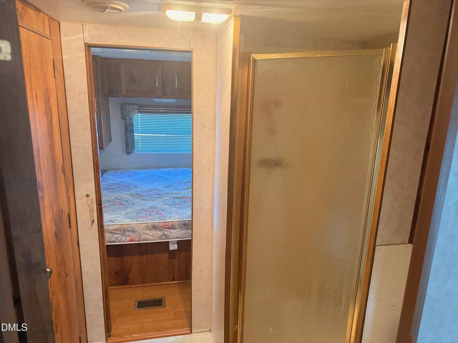 Interior view of an RV shows a frosted glass shower door on the right, wooden cabinets and a bed with a patterned bedspread in the background, and a vent on the wooden floor.