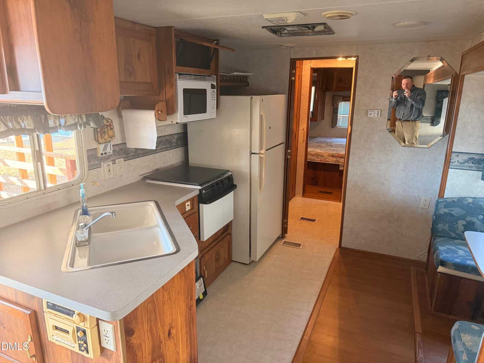 Interior view of an RV kitchen with a sink, stove, microwave, refrigerator, and dining area. A man is visible in the mirror taking a photo, and a bed is seen through a doorway in the back.