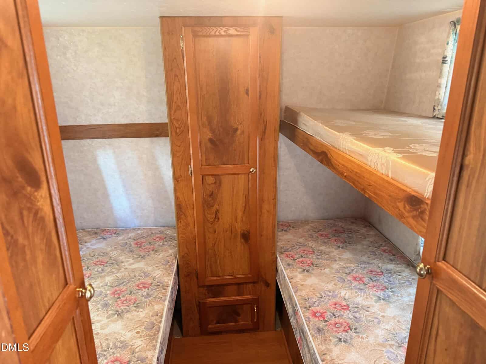 Small room with wooden walls and a central wooden cabinet, featuring three twin mattresses—two on the floor on each side and one upper bunk on the right. A window with a curtain lets in natural light.