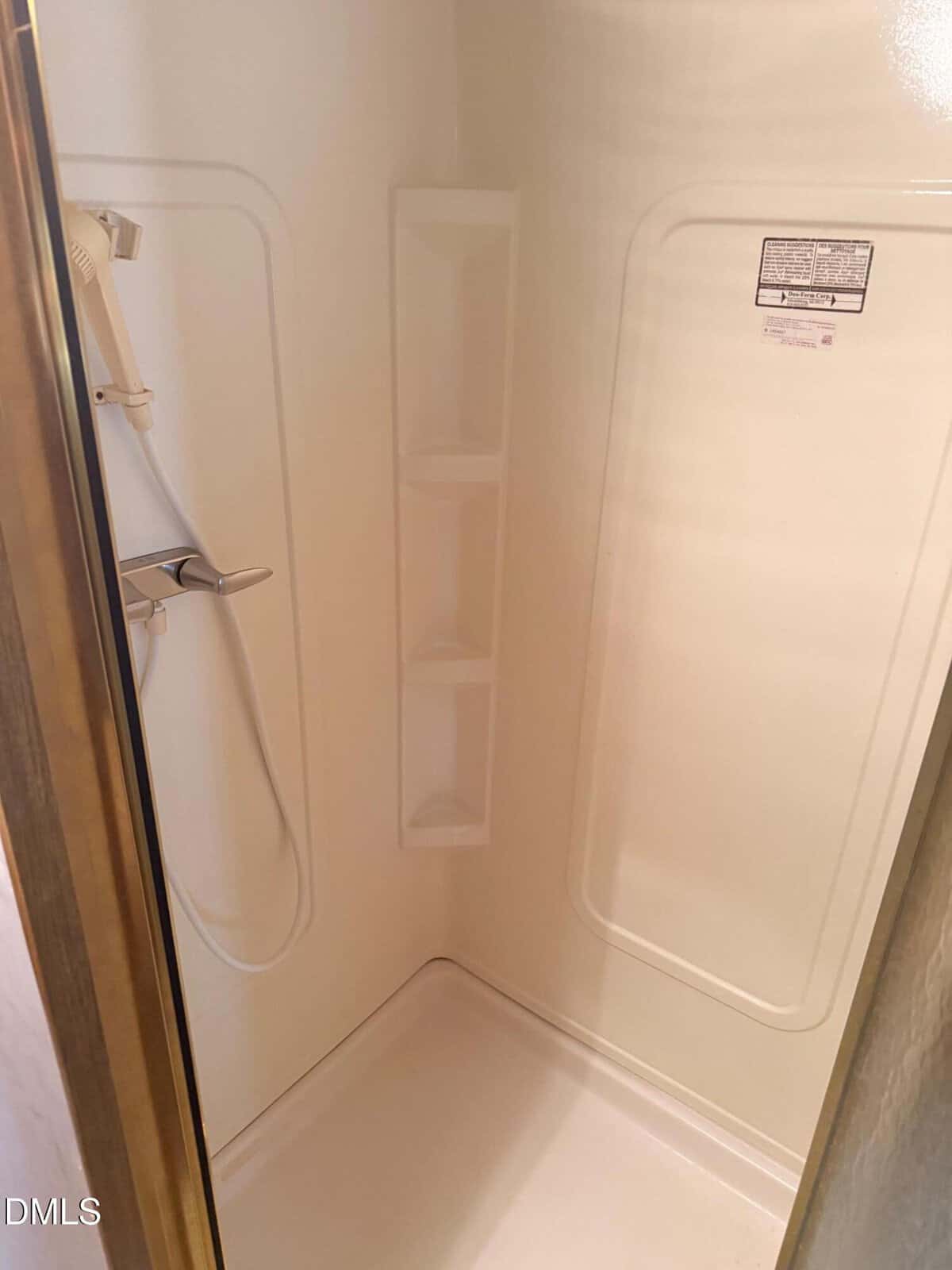 A clean, white shower stall with built-in corner shelves, a handheld showerhead, and a gold-colored frame partially visible on the left side.