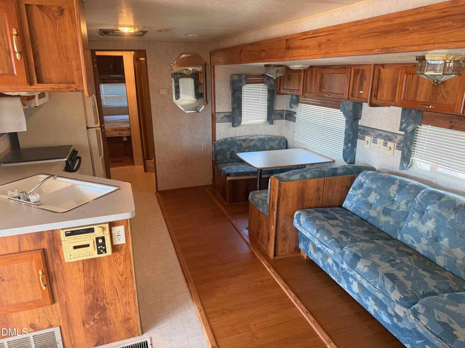 Interior of an RV with wood paneling, blue patterned sofa, matching dining booth, small kitchen with sink, stove, and fridge. Light streams through windows with blue valances. Mirror and hallway door at far end.