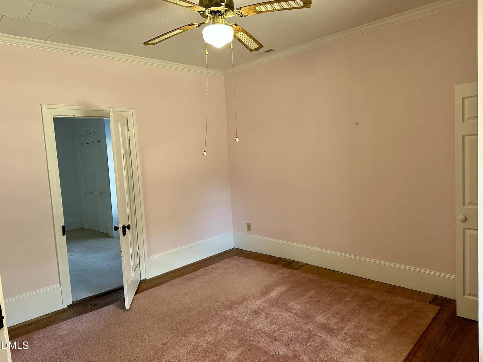 Empty room with pale pink walls, a ceiling fan with light, pink carpet, wooden floor trim, two white doors (one open to another room), and white crown molding.