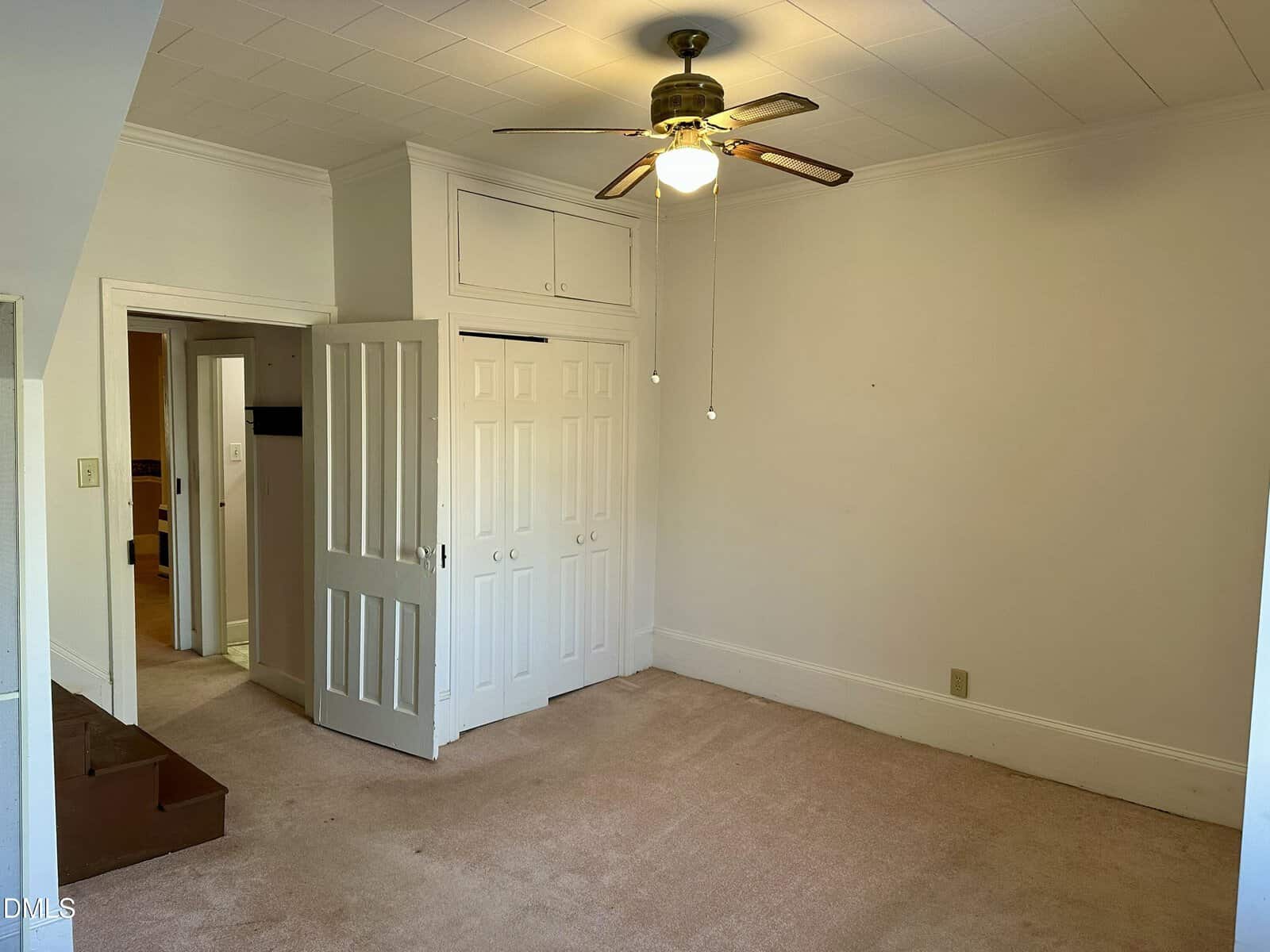 Empty room with beige carpet, white walls, a ceiling fan with light, open closet doors, a small staircase at left, and built-in storage above the closet.