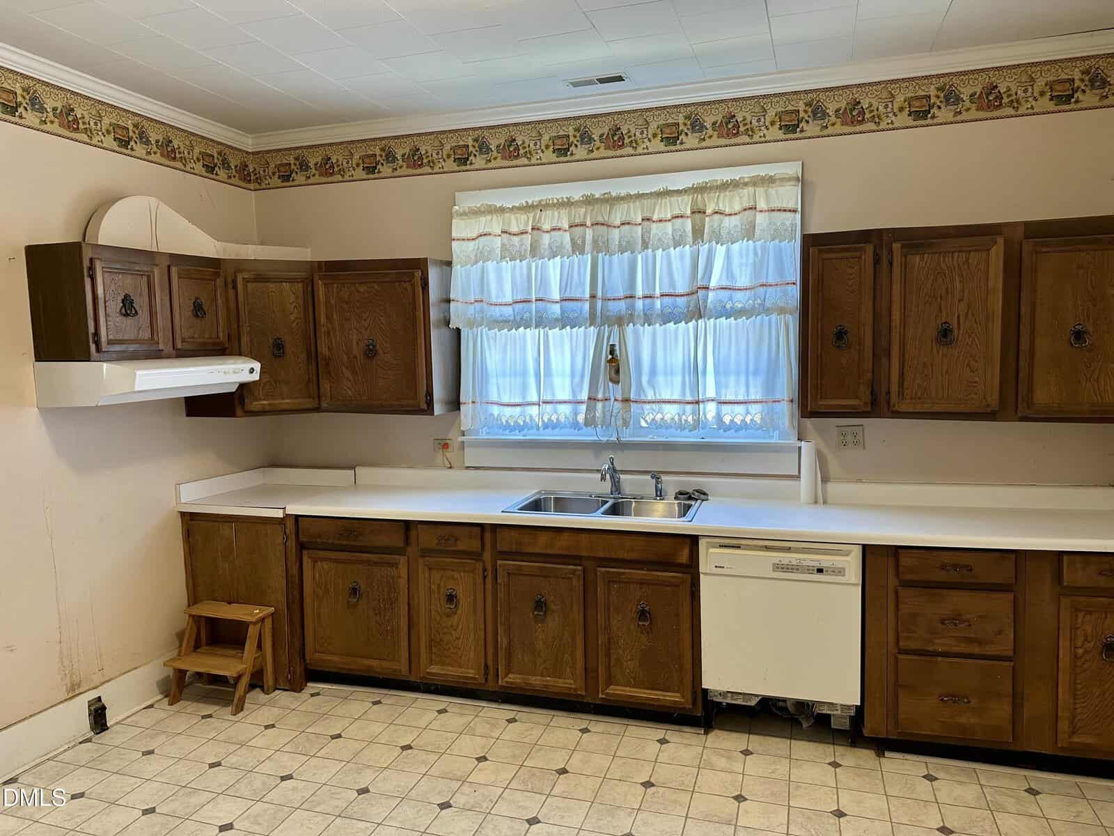 A vintage kitchen with wood cabinets, white countertops, a sink under a window with sheer curtains, a dishwasher, a white range hood, and a small wooden step stool on a tiled floor.