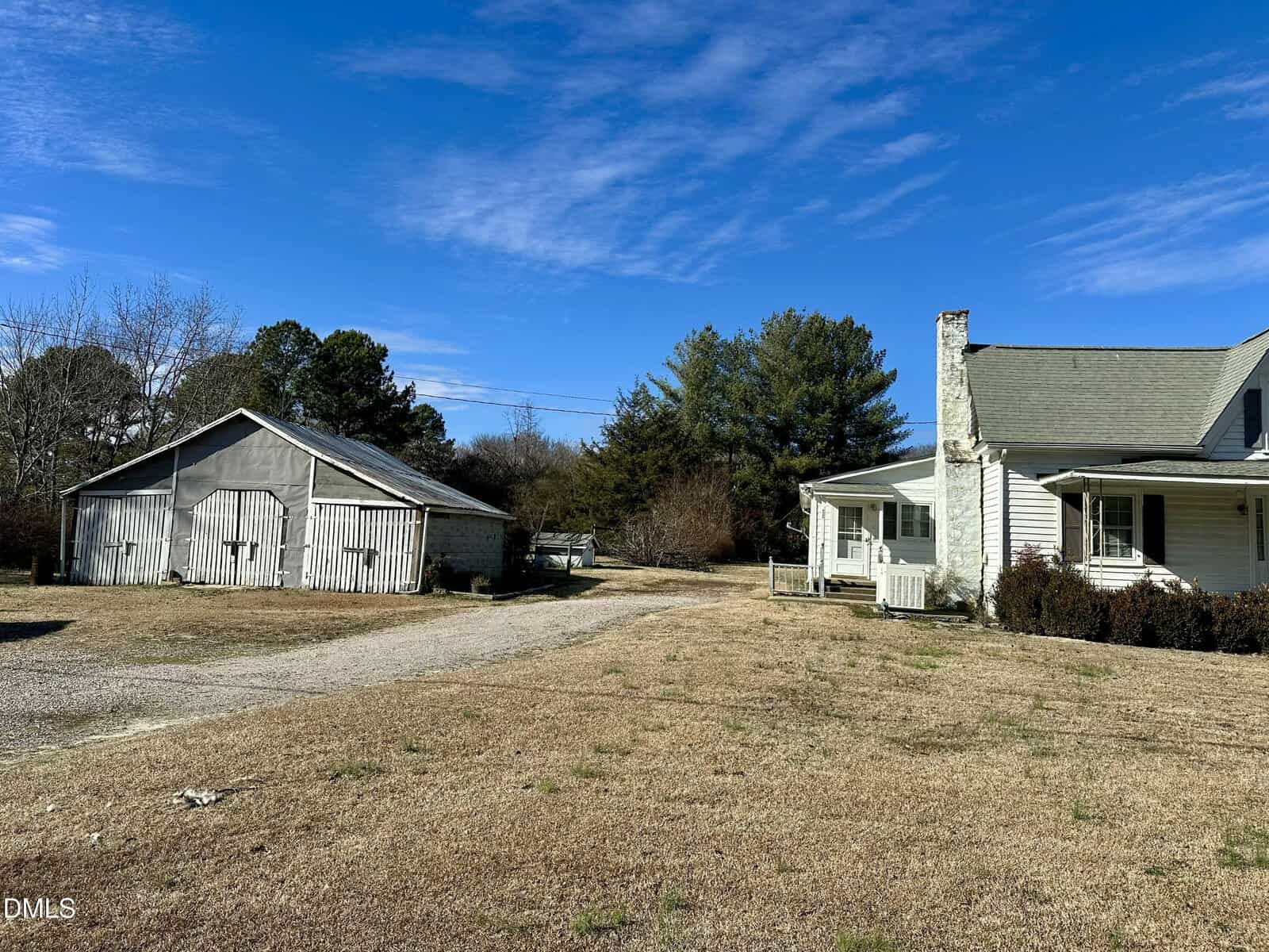 A white house with a porch sits on a dry, grassy yard next to a weathered gray garage. Trees border the property, and the sky above is clear and blue with a few wispy clouds.