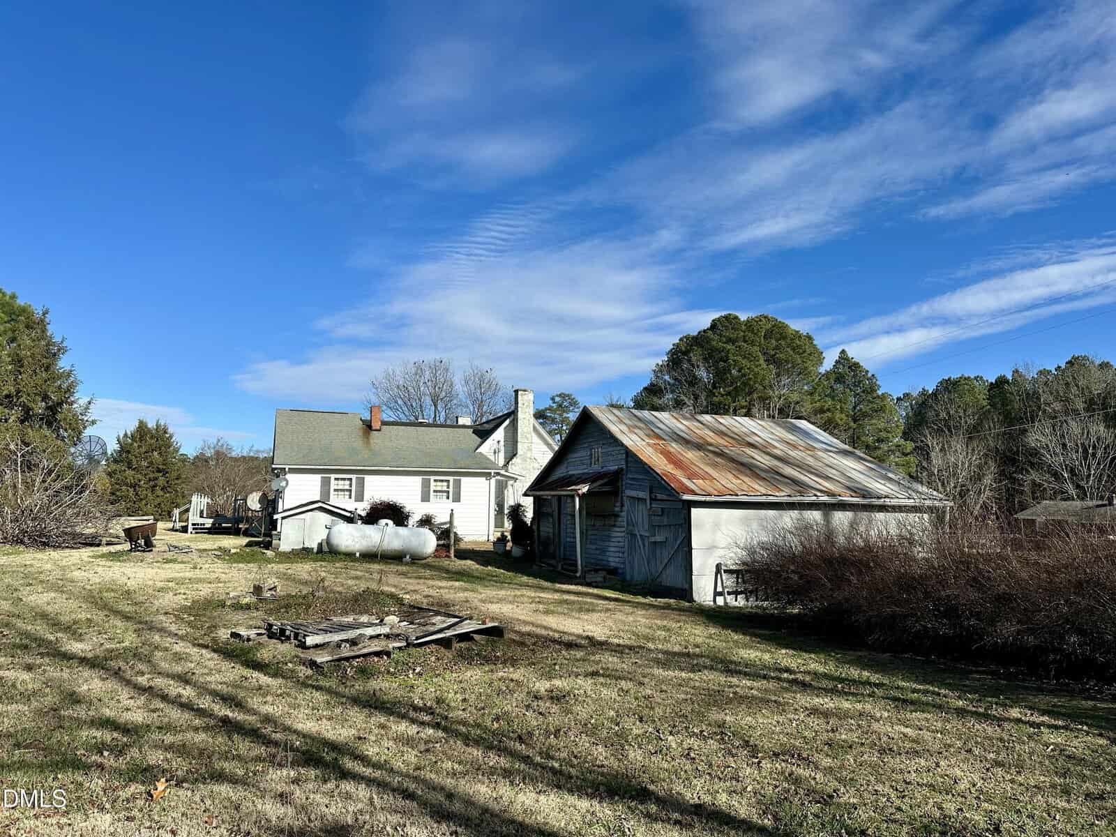 A small white house and an old, weathered barn with a rusted metal roof sit on a grassy lot under a blue sky with wispy clouds. Leafless trees and bushes surround the property.