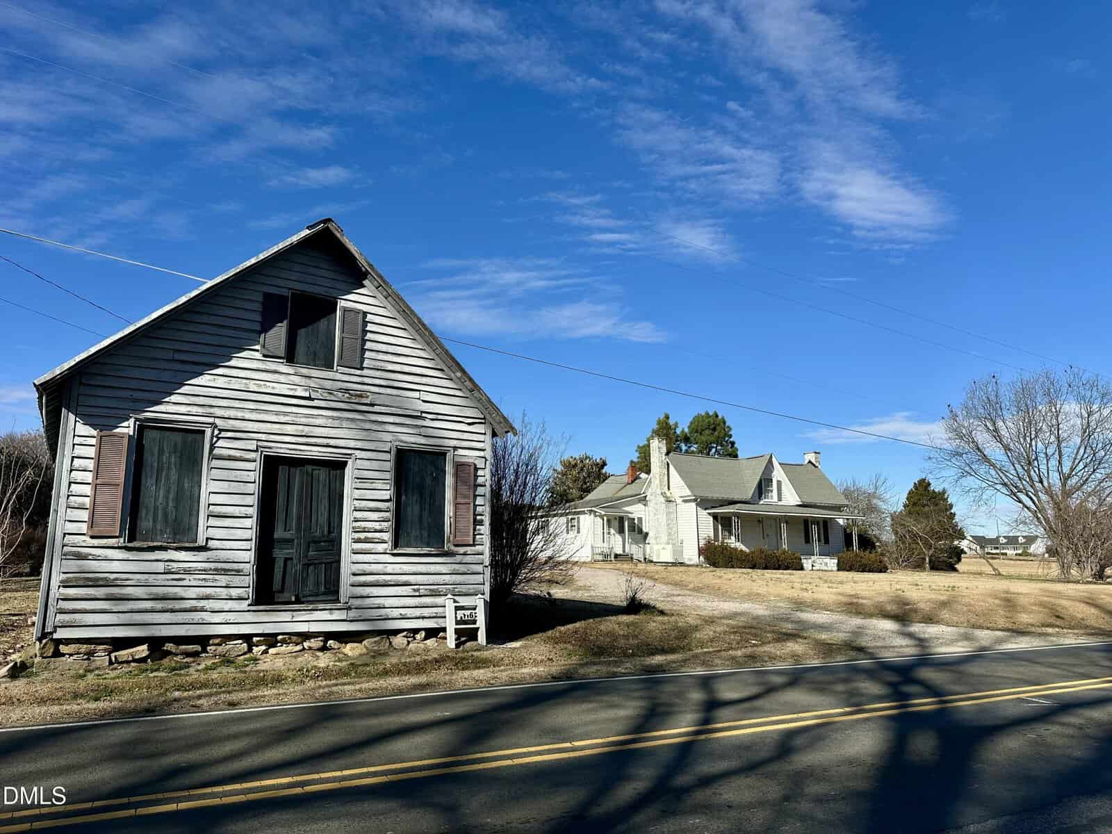 A weathered wooden building with shuttered windows sits by the roadside, with a larger white house and leafless trees in the background under a clear blue sky.