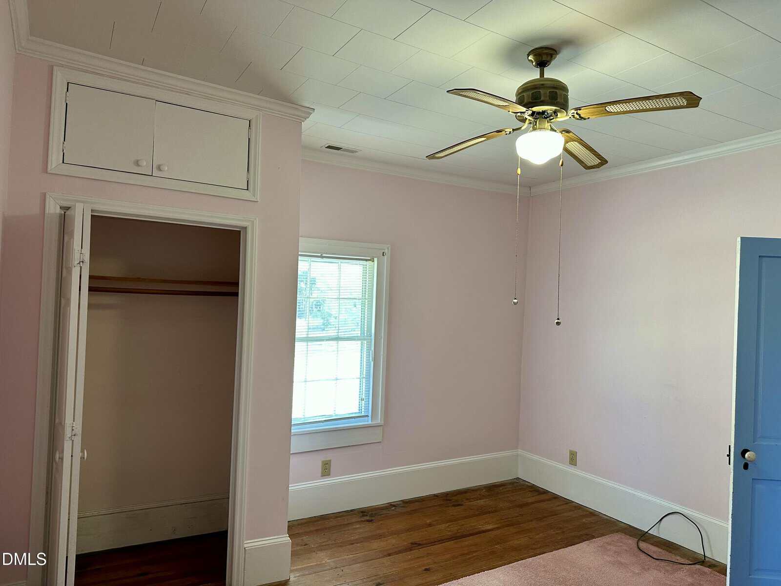 A small room with pink walls, wood floor, a ceiling fan with lights, an open closet, a window with blinds, and a blue door.