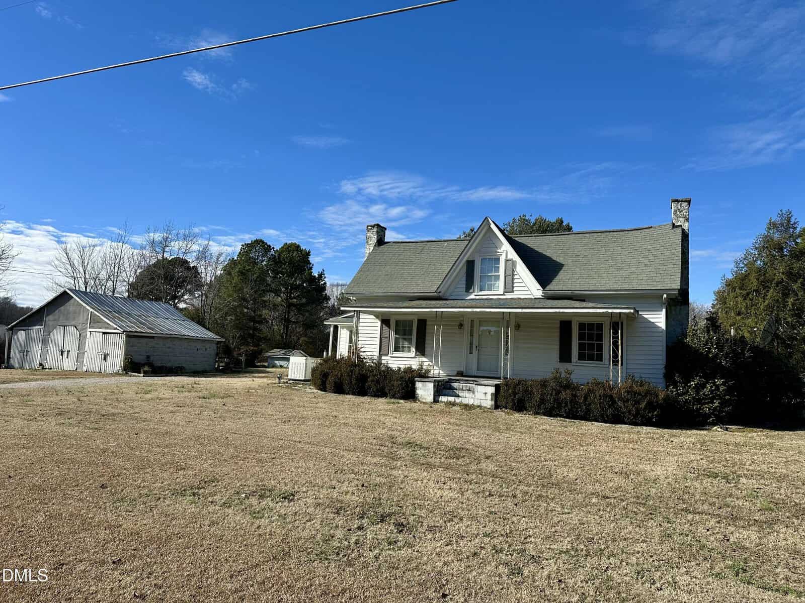 A white, two-story farmhouse with a covered porch and chimney sits on a large, grassy lot. An old barn and trees are visible in the background under a clear blue sky.