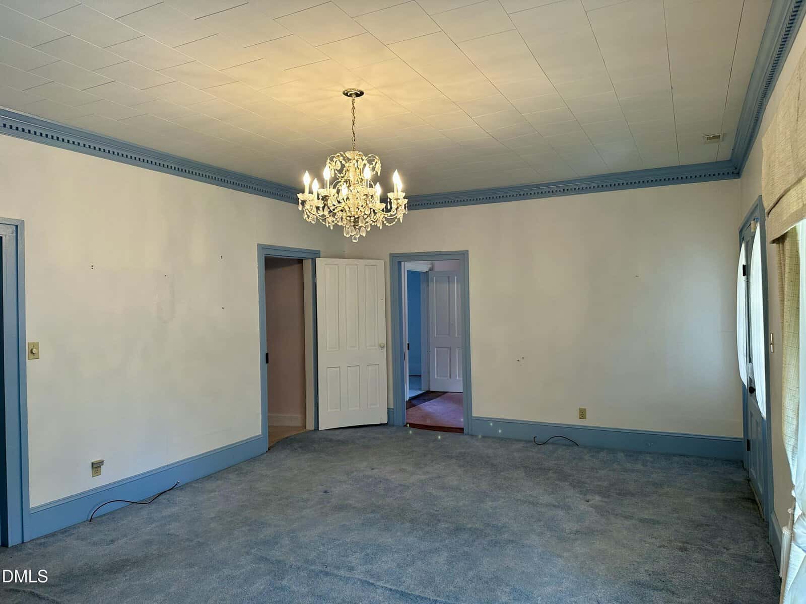 A mostly empty room with light blue carpet, white walls, blue trim, a chandelier ceiling light, three doorways, and large windows on the right letting in natural light.