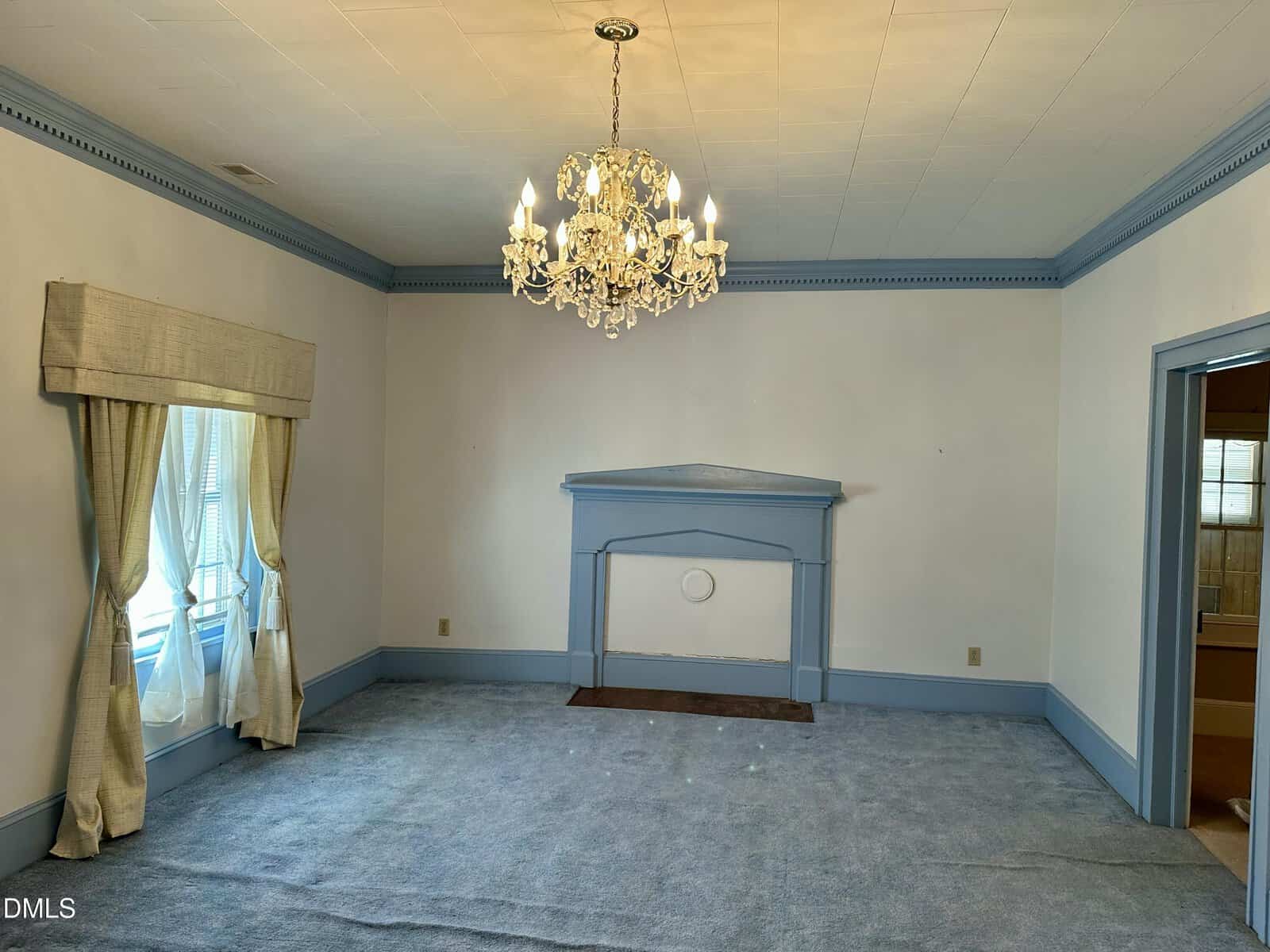 A mostly empty room with blue carpet, blue trim, a decorative blue mantel, white walls, a window with cream curtains, and a crystal chandelier hanging from the ceiling.