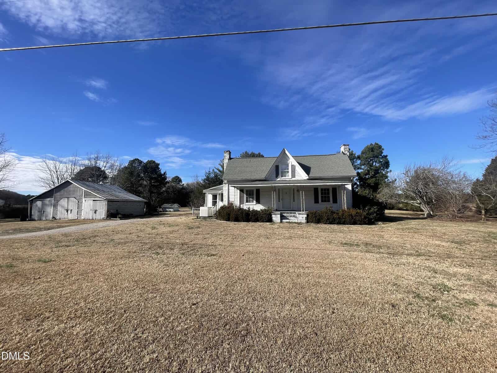 A white farmhouse with a covered front porch sits on a large, grassy lot under a blue sky. A barn is visible to the left, with trees surrounding the property and scattered clouds above.