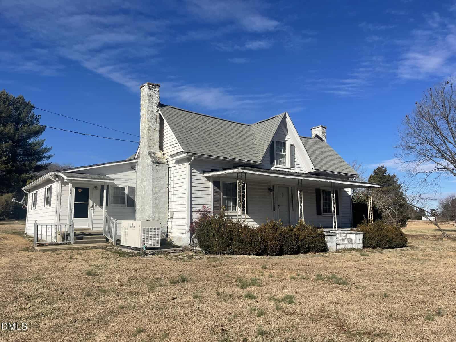 A white, older farmhouse with a covered front porch, stone chimney, and gabled roof sits on a large, dry grassy lot under a clear blue sky. Shrubs grow near the porch; trees are in the background.