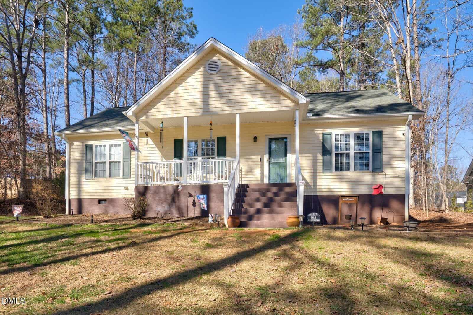 Charming single-story beige house at 103 Pueblo Drive, Louisburg, featuring a green door, white porch railings, front steps, and a sloped roof, nestled among trees and a lush lawn on a sunny day.