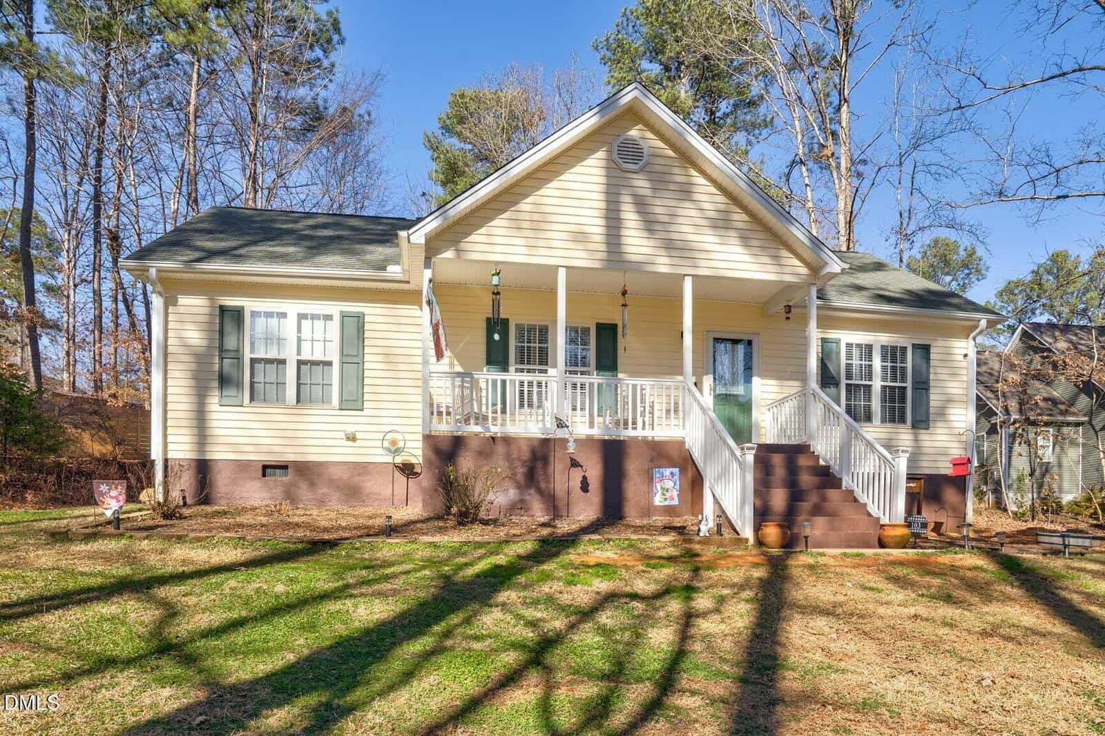 A beige single-story house with white trim, a covered front porch, green shutters, and a green front door. Leafless trees and shadows cover the lawn on a sunny day.