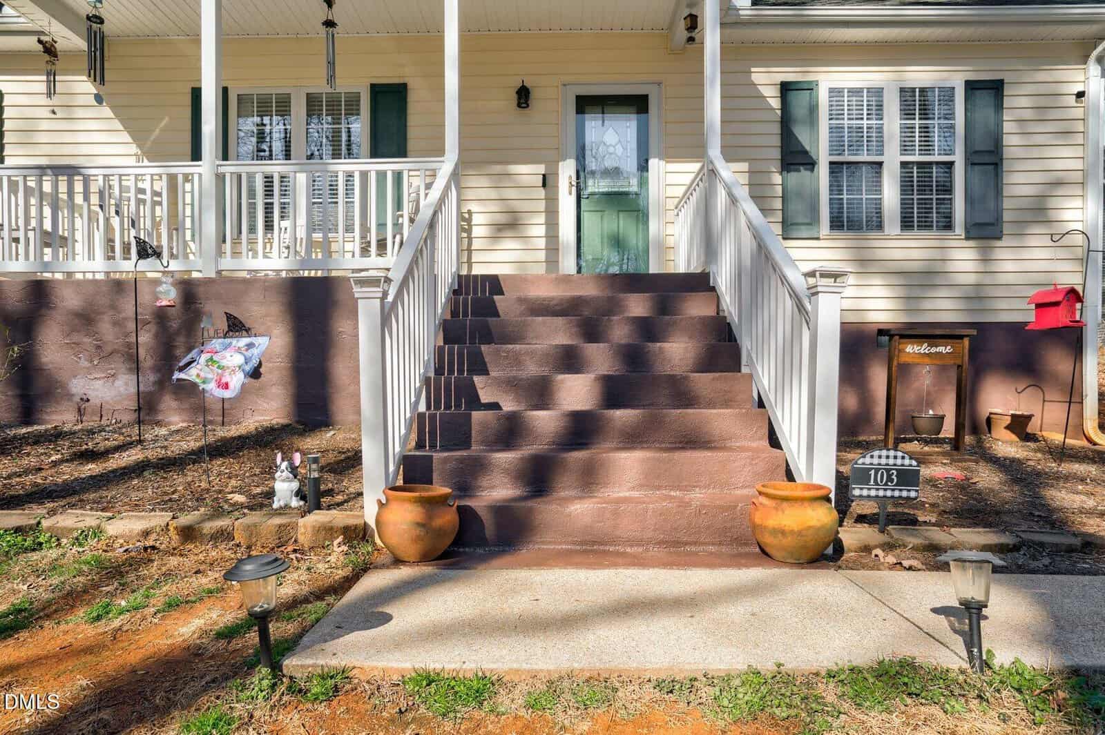 Front porch with brown steps, white railings, two large terracotta pots, a “Welcome” sign, house number 103, and decorative items on soil and mulch areas near the stairs.
