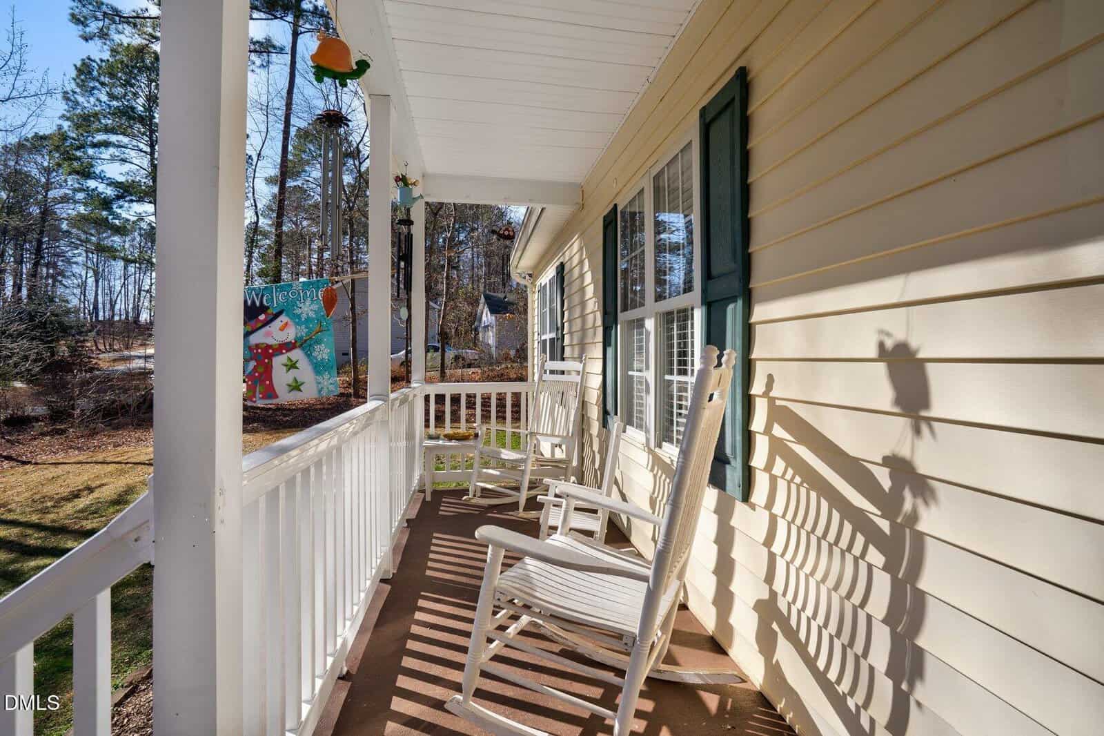 A cozy front porch with white railings and rocking chairs, attached to a yellow house with green shutters. A colorful "Welcome" flag and trees are visible in the background.