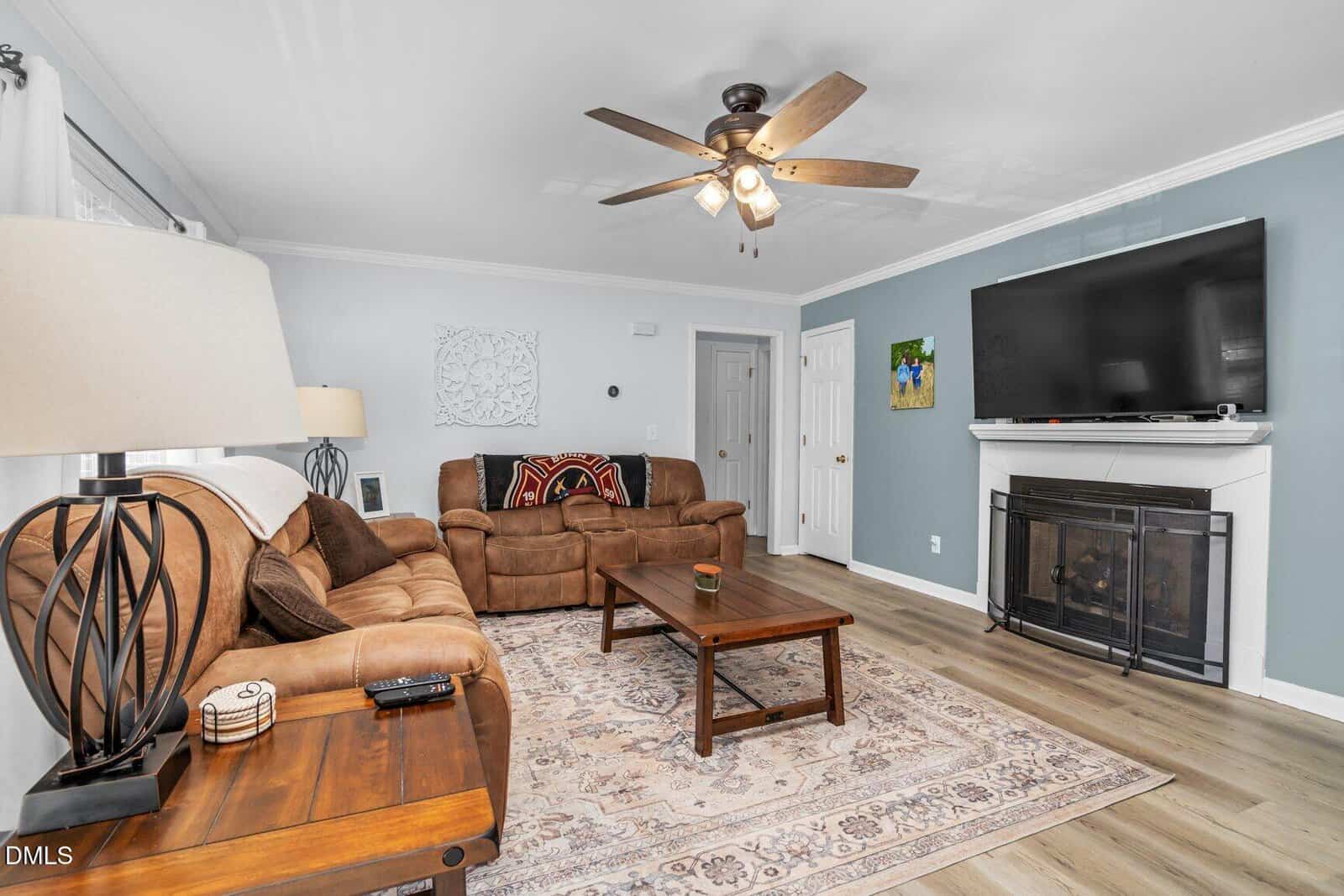 A cozy living room with brown couches, a wooden coffee table, a decorative area rug, a ceiling fan with lights, a wall-mounted TV above a fireplace, and light blue accent walls.