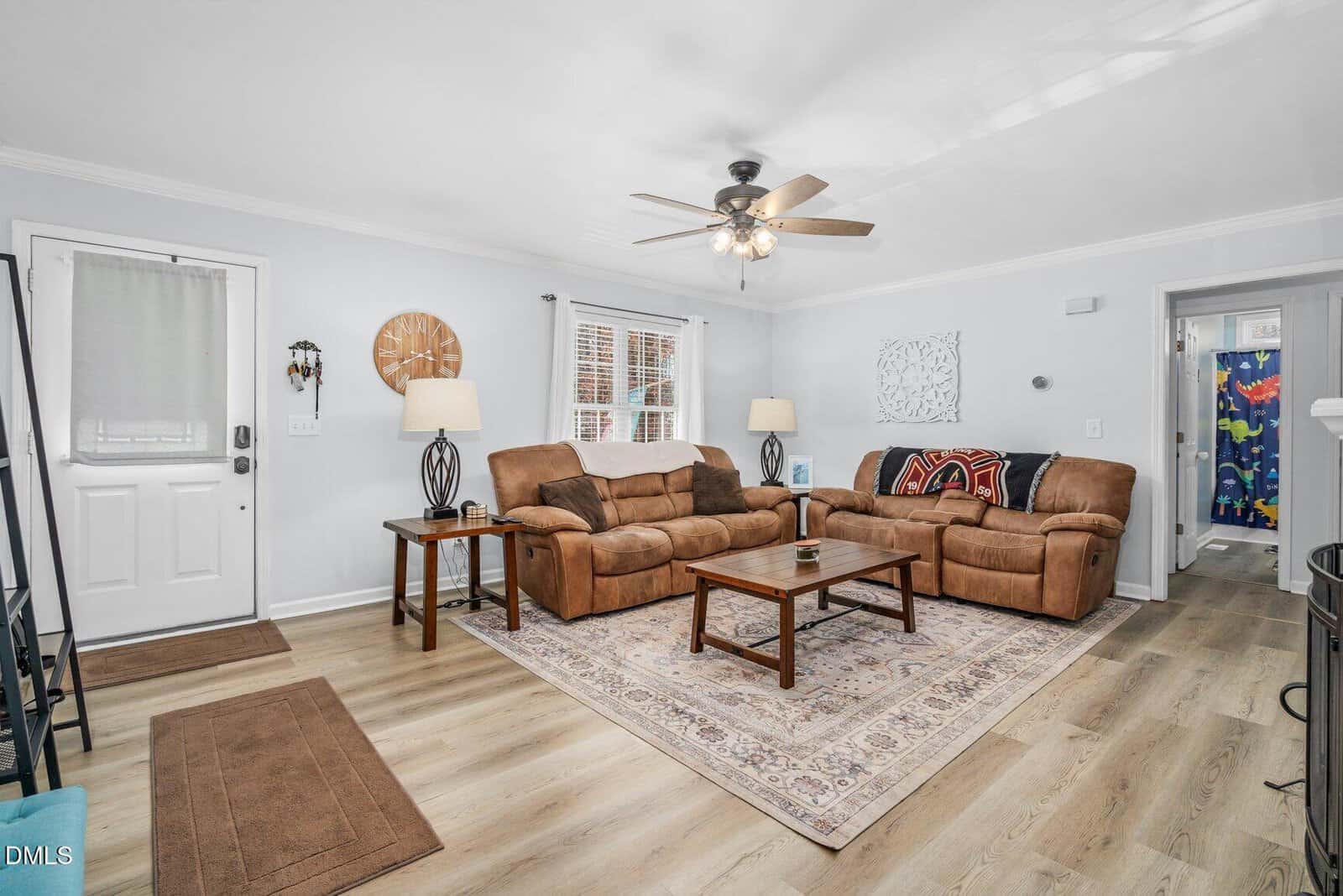 A cozy living room with two brown sofas, a wooden coffee table, two end tables with lamps, a rug, wall art, a ceiling fan, and light wood floors. A door and a window with blinds are on the left. A hallway is visible on the right.