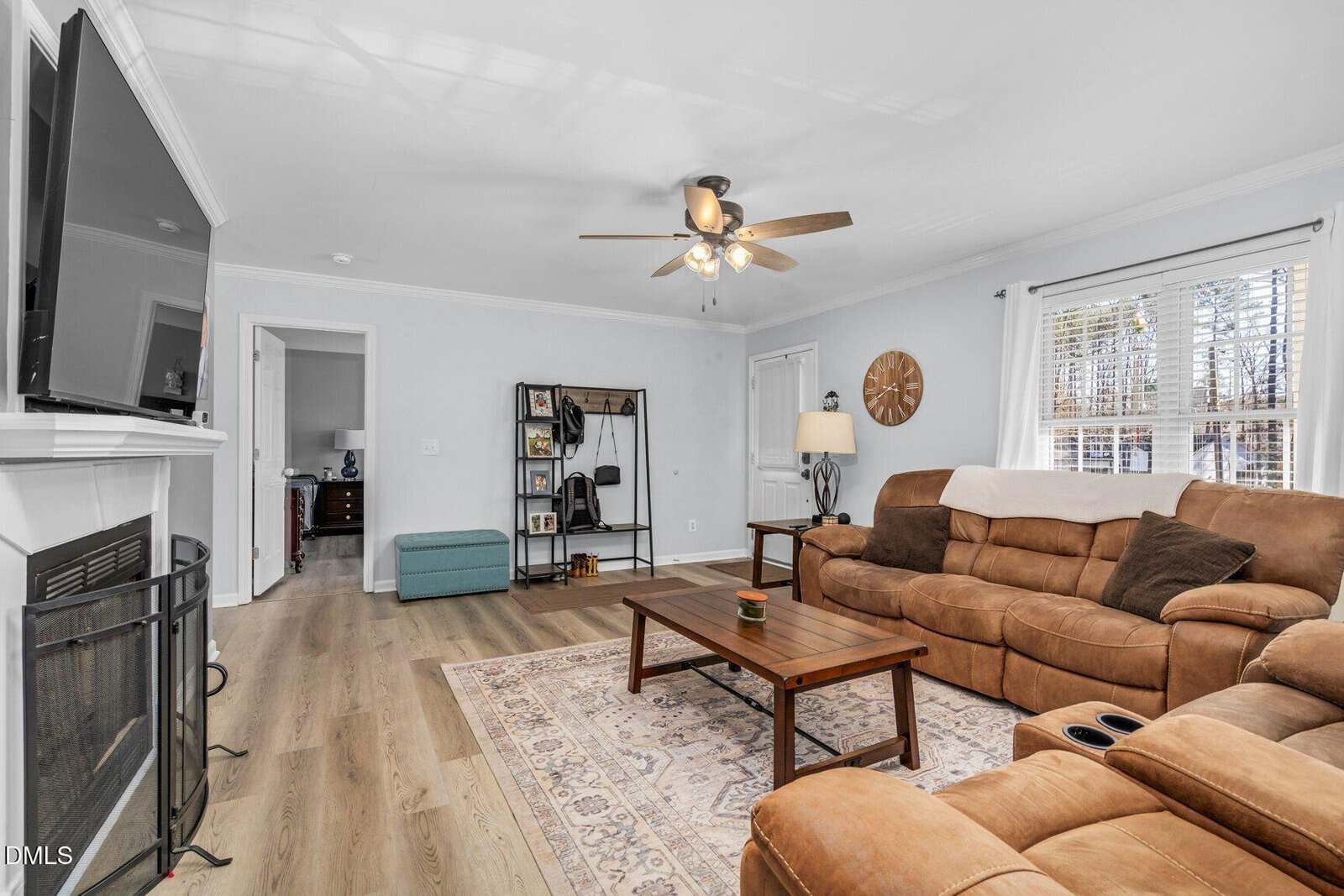 A cozy living room with a brown sectional sofa, wooden coffee table, ceiling fan, wall-mounted TV above a fireplace, and large window with blinds. Light wood flooring and a patterned rug complete the space.