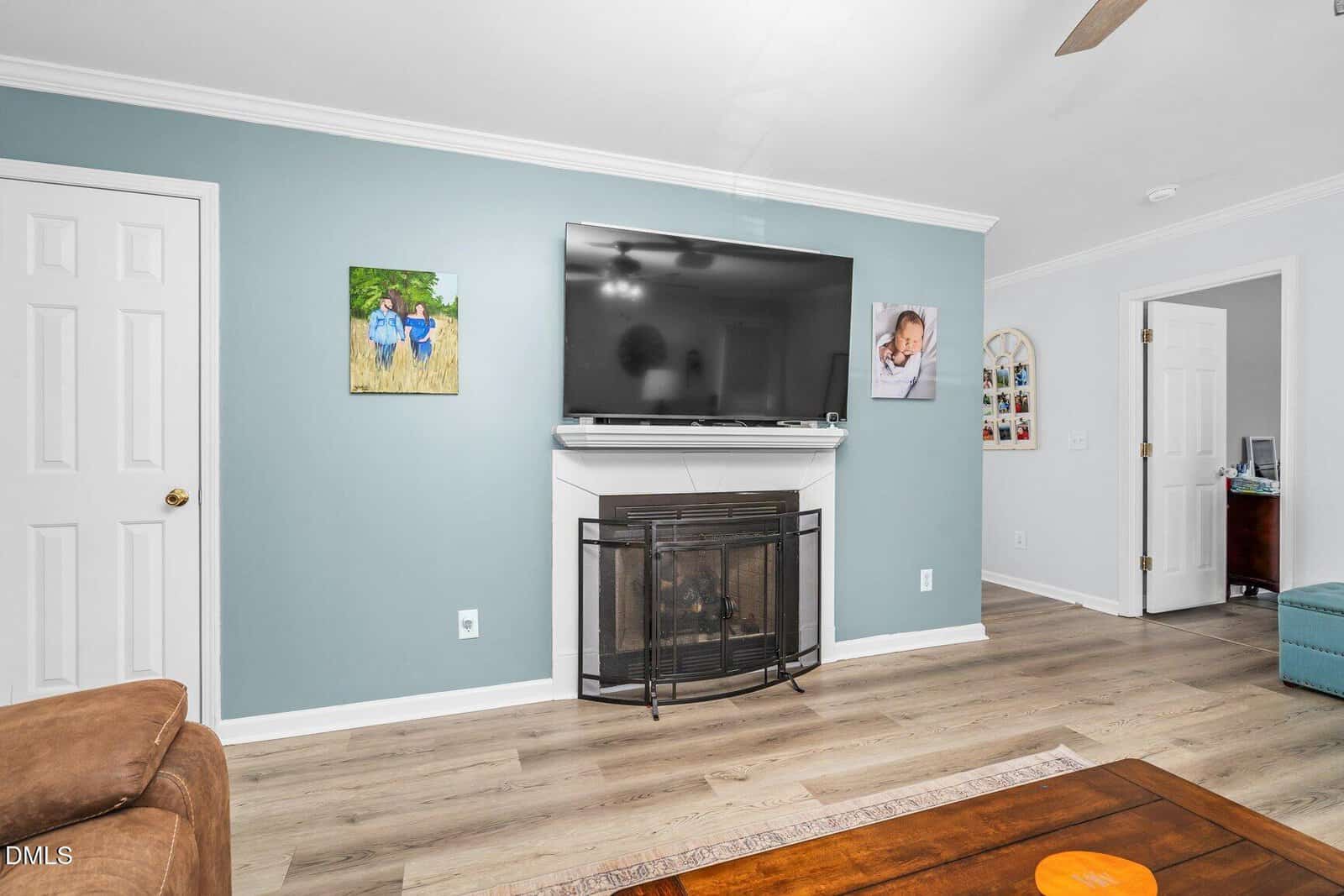 A living room with light blue walls, a wall-mounted TV above a corner fireplace, family photos on the wall, light wood flooring, and white doors. A brown couch and wooden coffee table are partially visible.