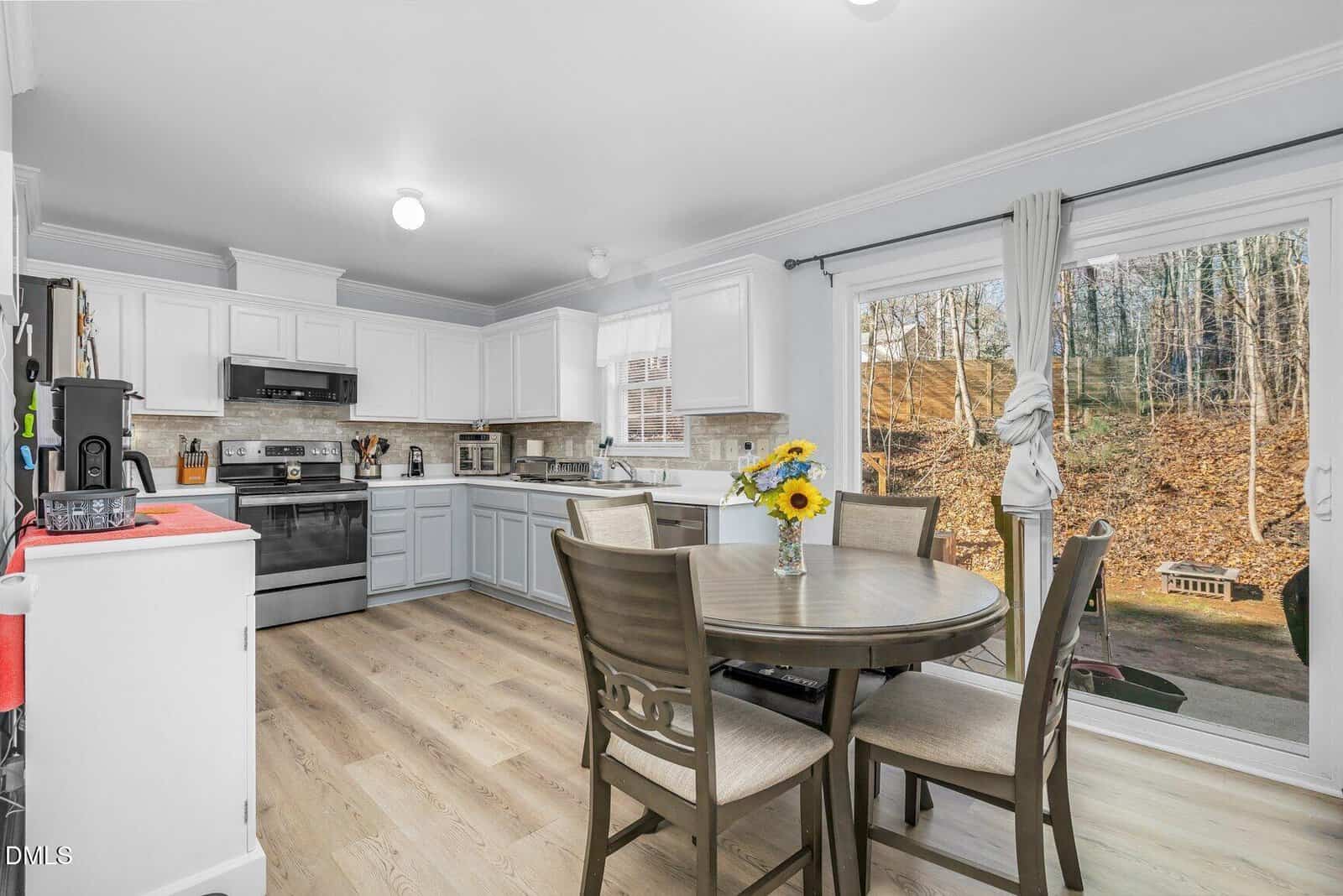 Bright kitchen with white cabinets, stainless steel appliances, and light wood flooring. A round dining table with four chairs and a vase of sunflowers sits near sliding glass doors overlooking a wooded backyard.
