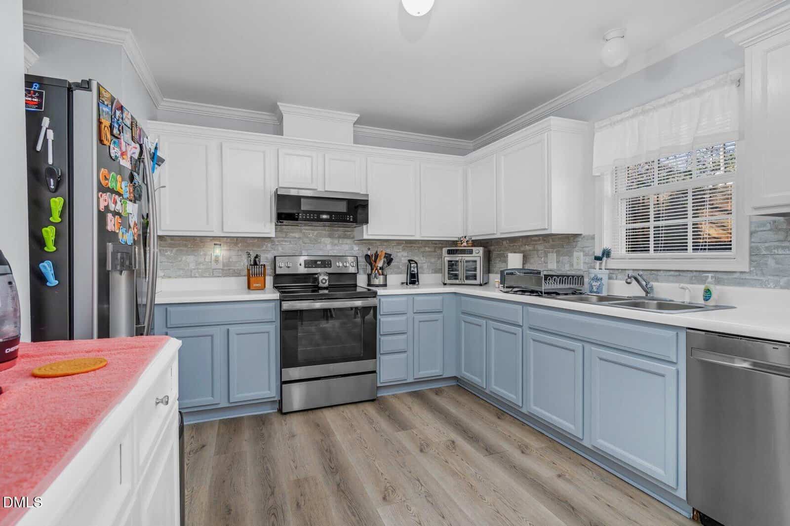 A modern kitchen with white upper cabinets, light blue lower cabinets, stainless steel appliances, and wood flooring. The refrigerator is decorated with colorful magnets, and light streams in through a window above the sink.