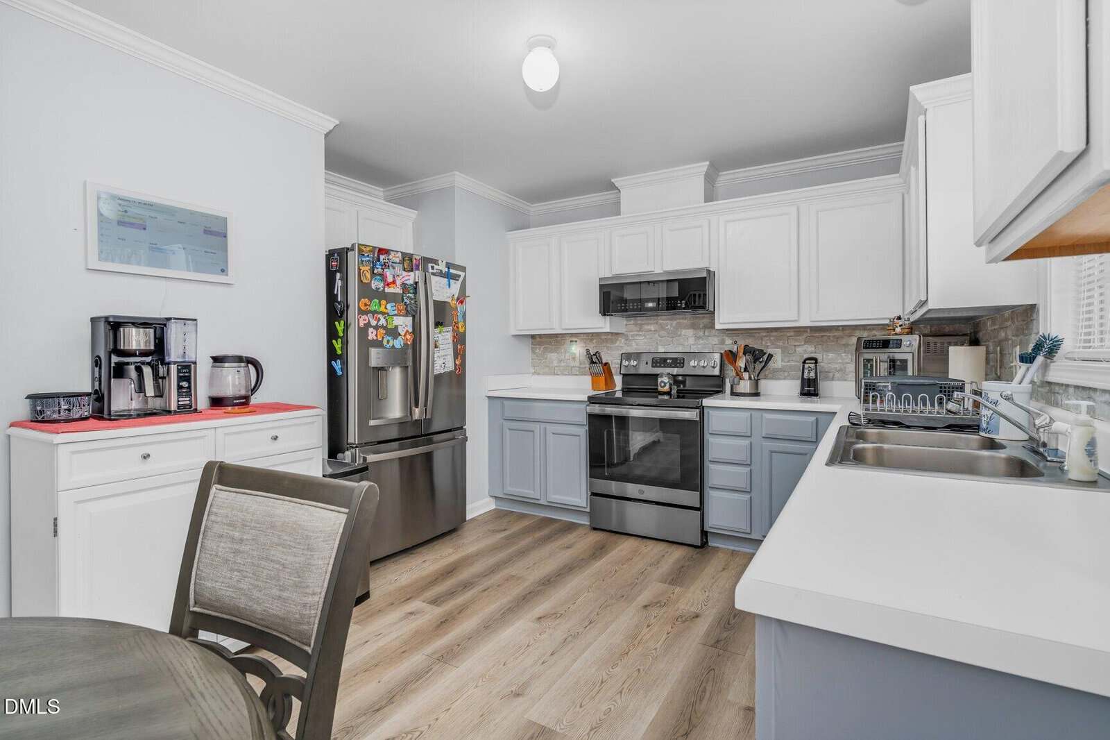 Modern kitchen with white and gray cabinets, stainless steel appliances, a refrigerator with magnets, coffee station, dining table, and wood-style flooring. Bright lighting and countertops with various kitchen items visible.