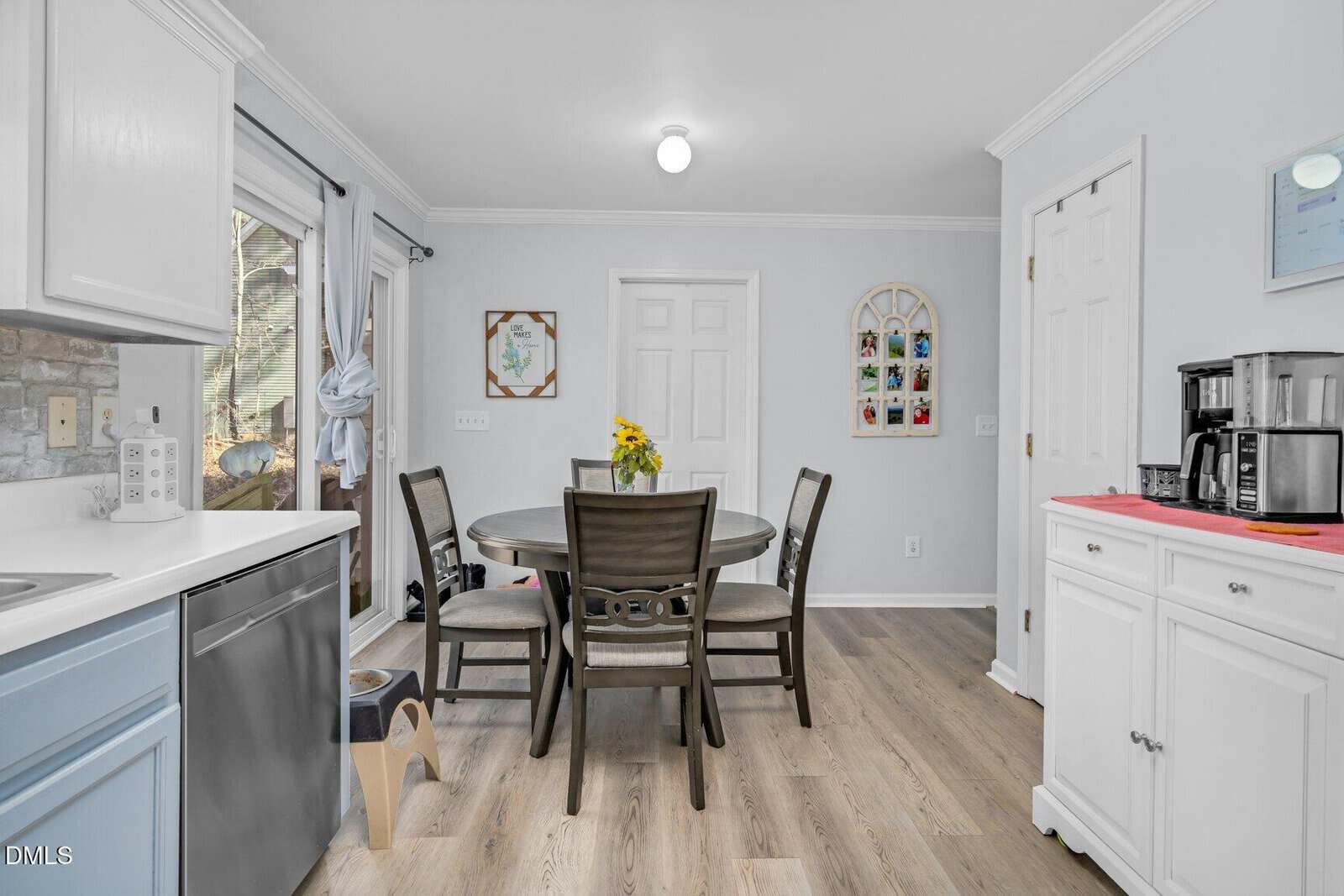 Bright kitchen with light wood floors, a round dining table with four chairs, white cabinets, coffee maker, art on the walls, and a window with light curtains letting in natural light.