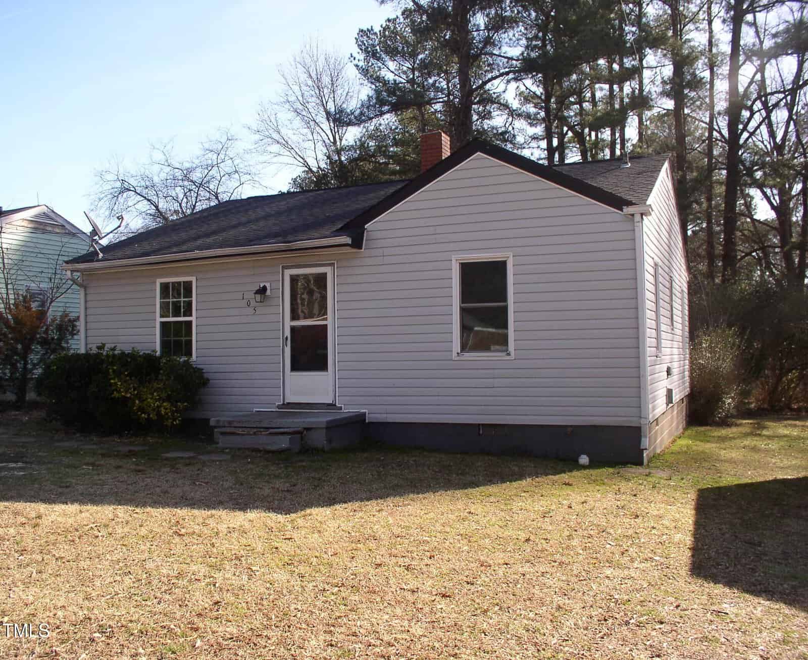 A small, single-story house at 105 S Elizabeth Street in Henderson features white siding, a dark shingle roof, and a brick chimney; two front windows frame the central door with small steps, set amid a grassy yard with trees and shrubs.