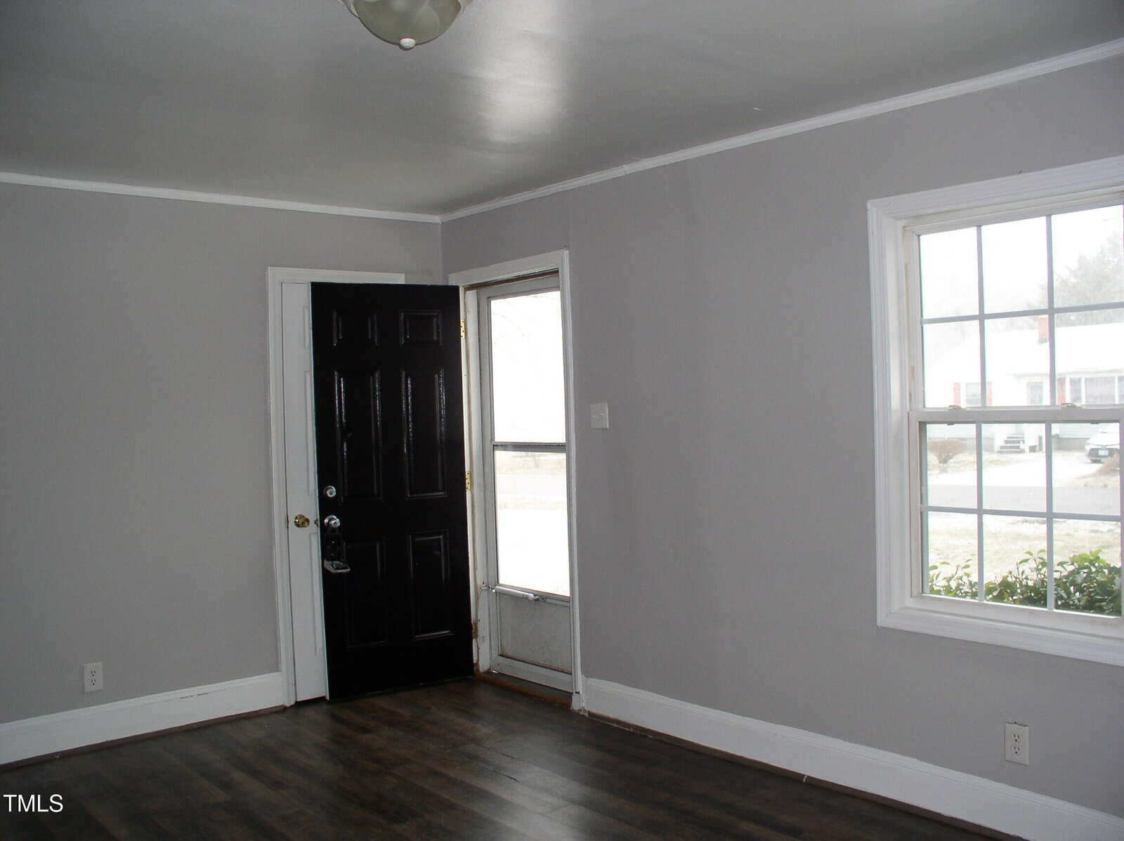 A small, empty room with gray walls, white trim, dark wood floors, a black front door, and a window letting in natural light.