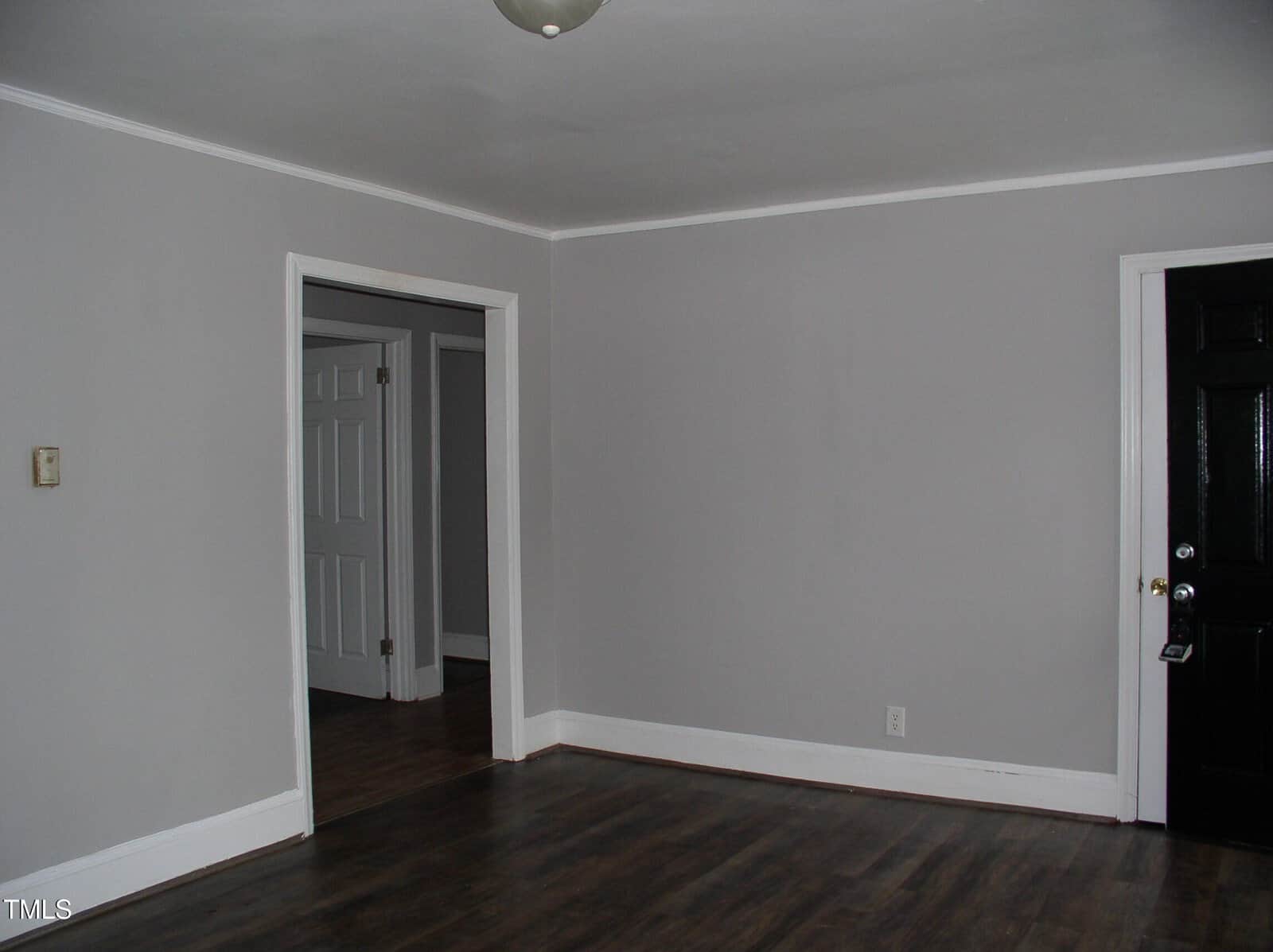 Empty room with gray walls, dark wood flooring, white trim, a black door on the right, and a doorway leading to another room with white doors. Ceiling light fixture is visible.