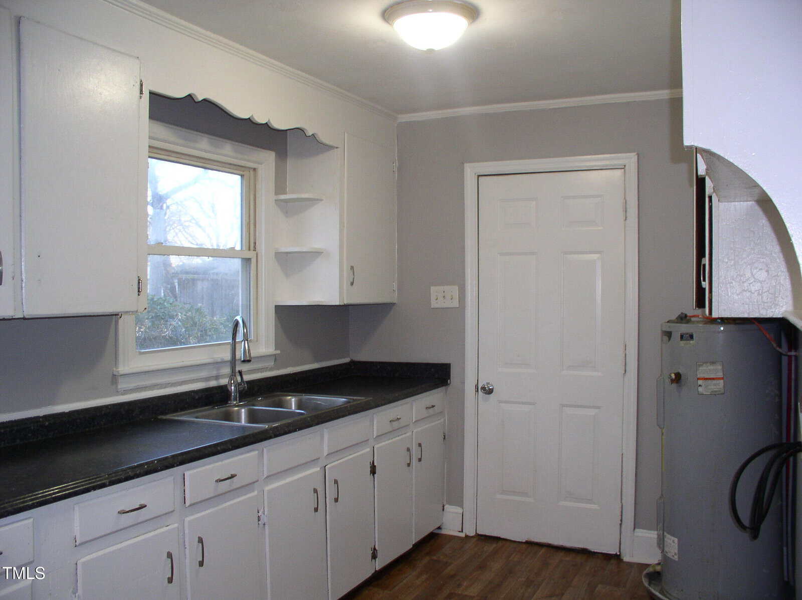 A kitchen with white cabinets, a double sink under a window, black countertops, wood flooring, gray walls, and a white door. A water heater is visible in the corner. Ceiling light is on.