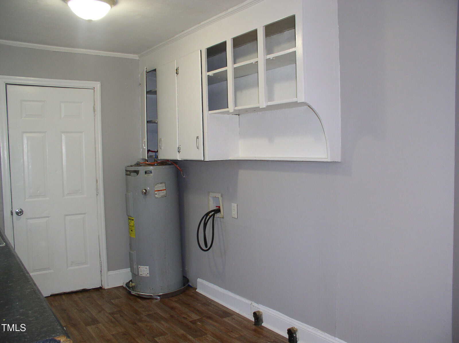 A utility room with light gray walls, wood flooring, a water heater, exposed hoses and pipes, white upper cabinets with glass doors, and a closed white door.